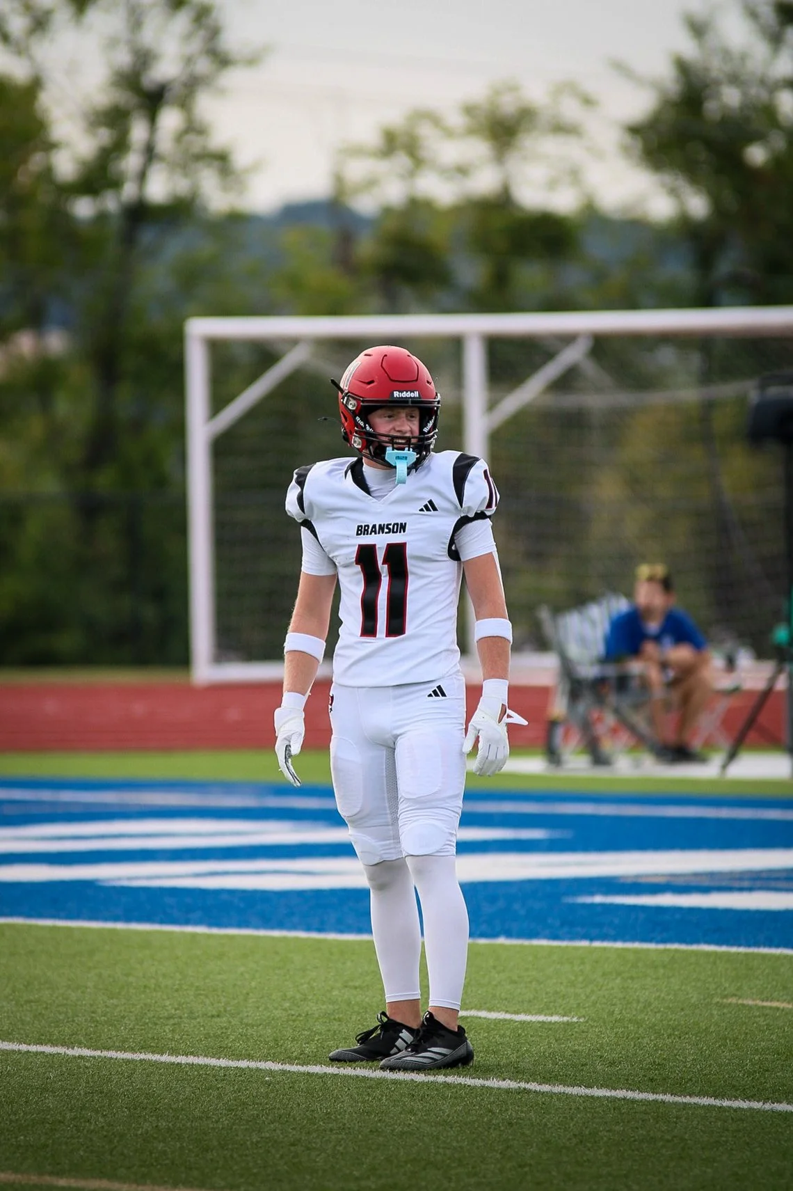Football player in a white uniform with the number 11 stands on a football field wearing a red helmet and gloves, with a goalpost in the background.