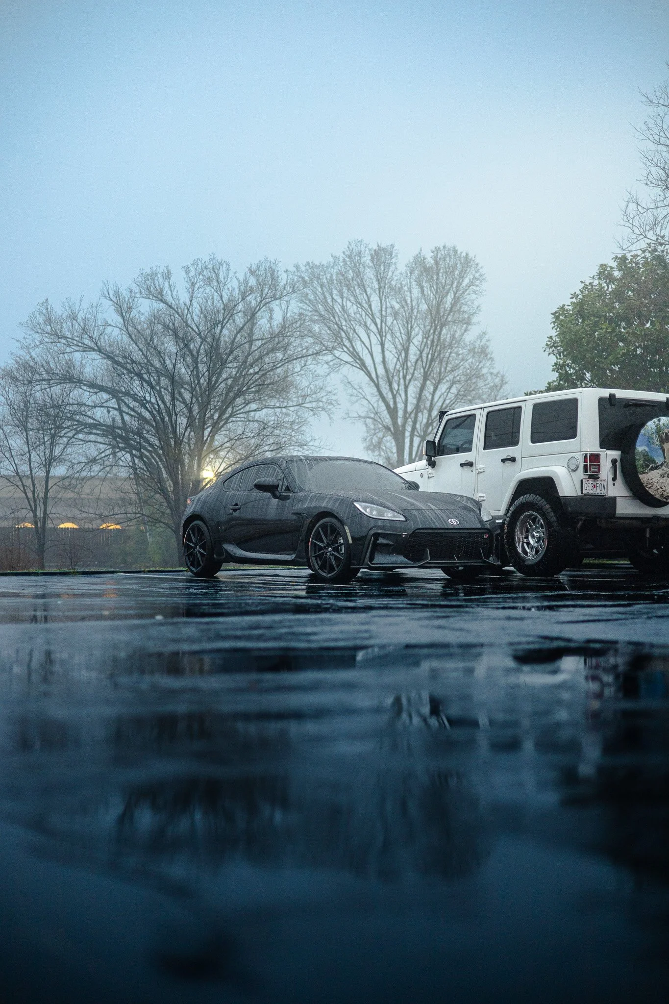 A black sports car and a white SUV parked on a wet parking lot with leafless trees and fog in the background.