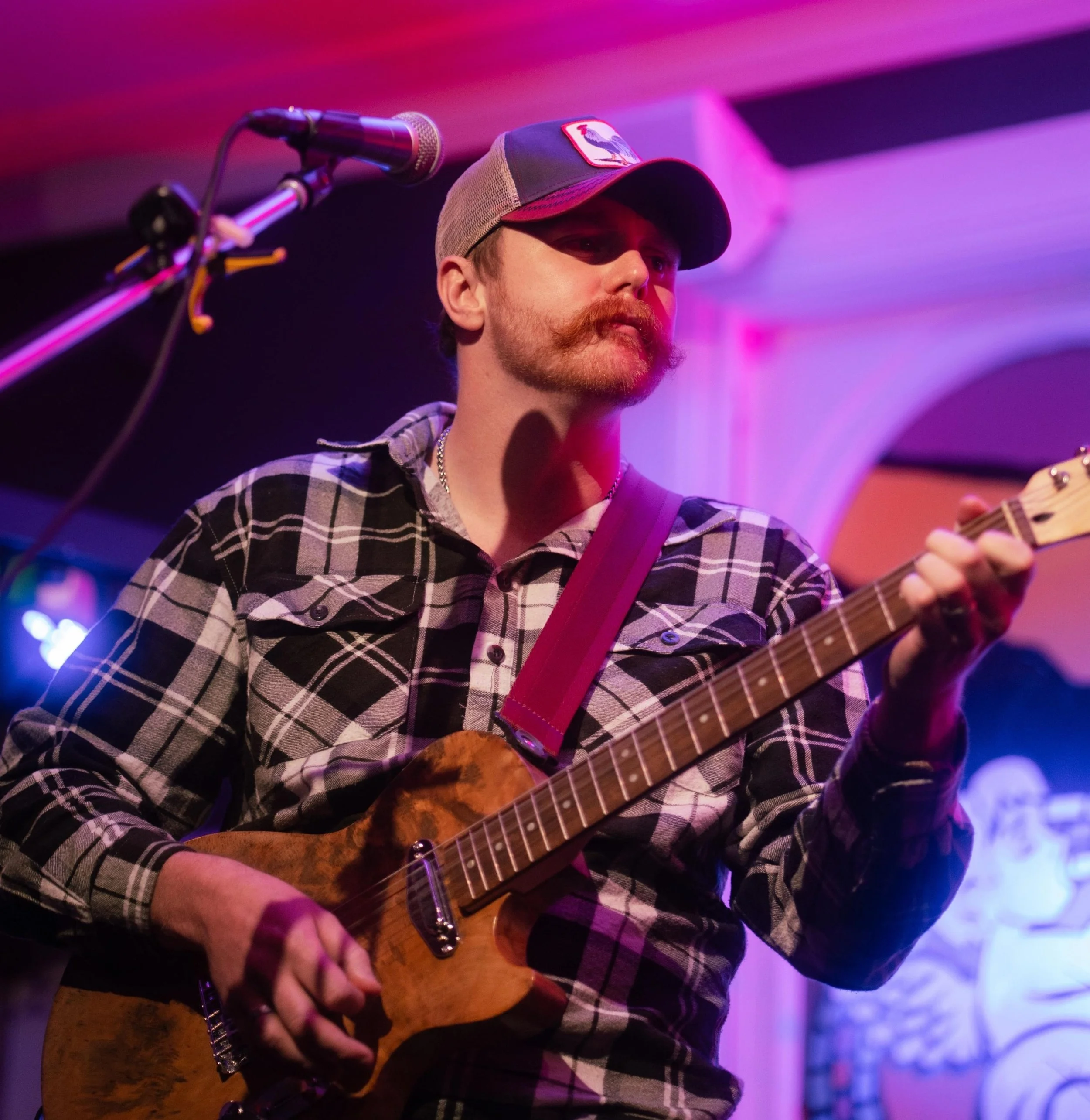 A man with a red beard and mustache, wearing a baseball cap and plaid shirt, playing an acoustic guitar on stage with colorful lighting.
