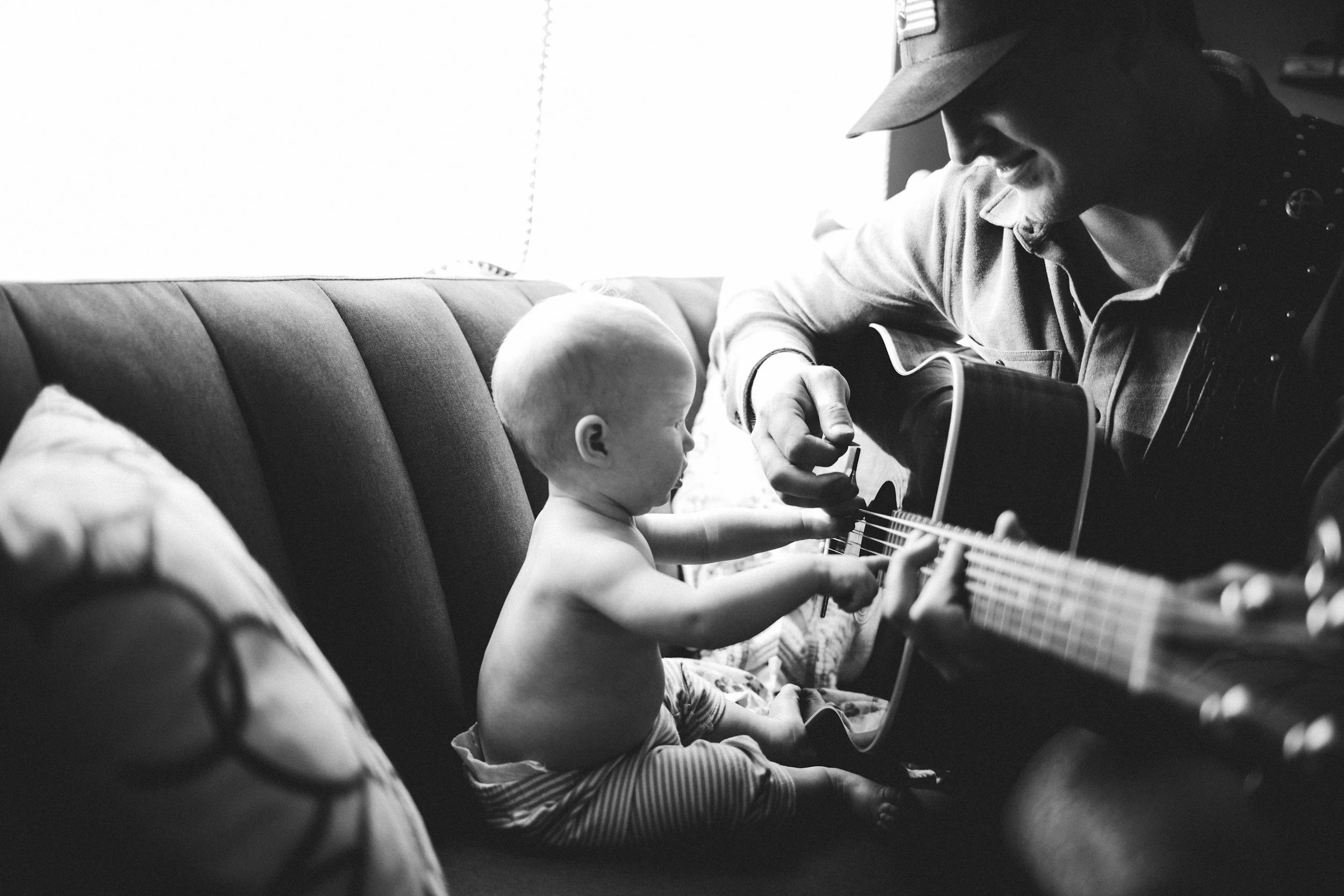A man playing a guitar for a young child, likely his son, who is sitting on a couch, as the child reaches for the guitar strings. Demonstrates the importance of fathers in their children's lives and of fatherhood for men.