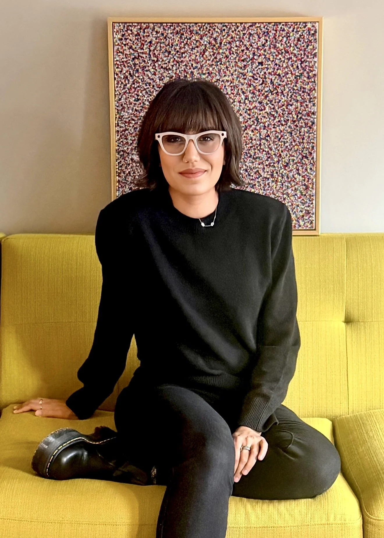 Artist Ashley Strayhorn of Artwork by AMS. Ashley wears a black sweater and glasses, sitting on a bright yellow couch. Behind her, a large square sculptural panel of multicolored glass rhinestones is displayed in a gold frame on a gray wall.
