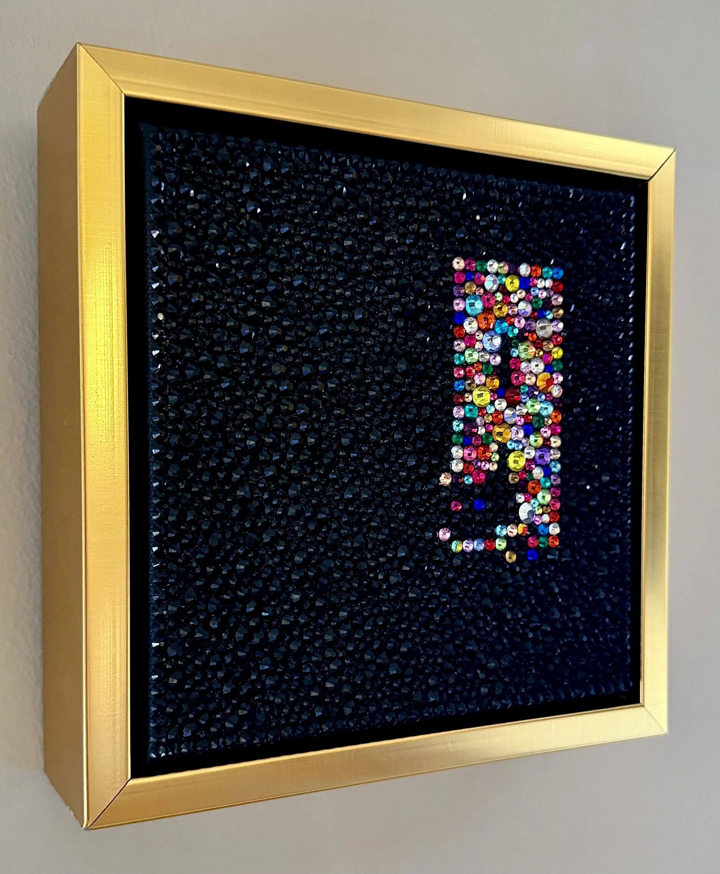 Angled side view of "Unease," showing the depth of the black rhinestone texture and the inset multi-colored rhinestone cluster within a gold floating frame.