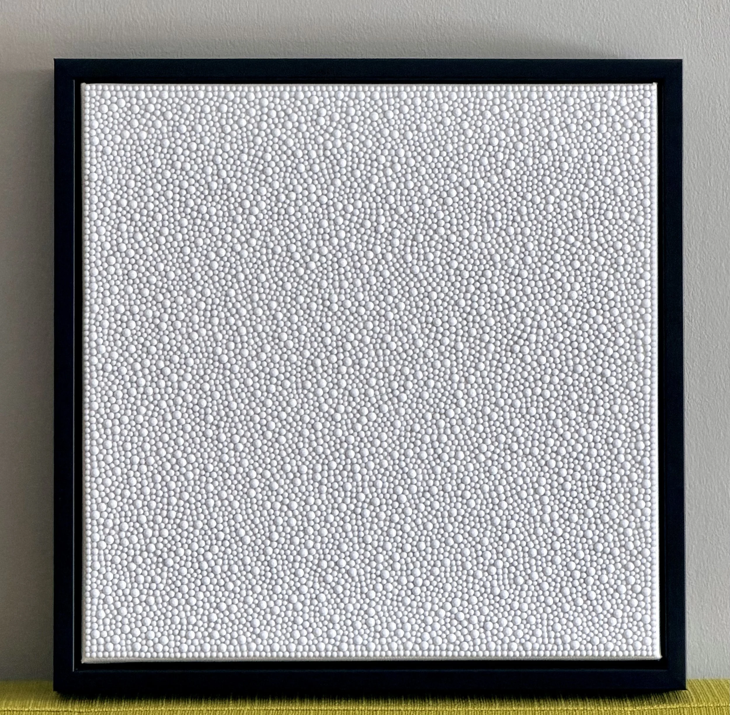 Flawless (2025) displayed on a green couch. The 12x12 in. white sculptural panel’s brilliant shimmer and 3D texture stand out against the bright green.