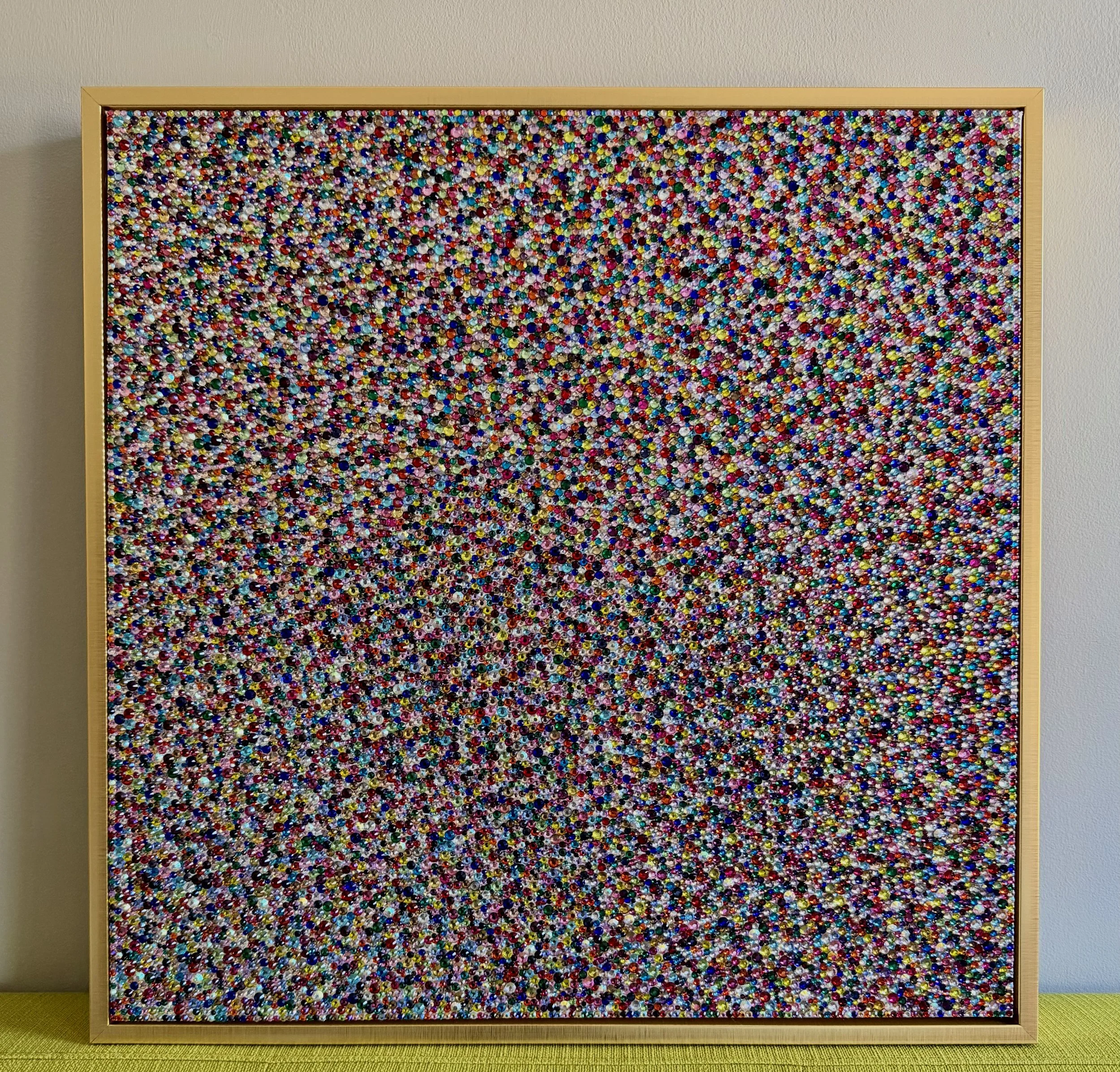 Panic (2026), 20x20 in. Multicolored sculptural panel in a gold frame, displayed on a yellow couch to show scale and prismatic texture.
