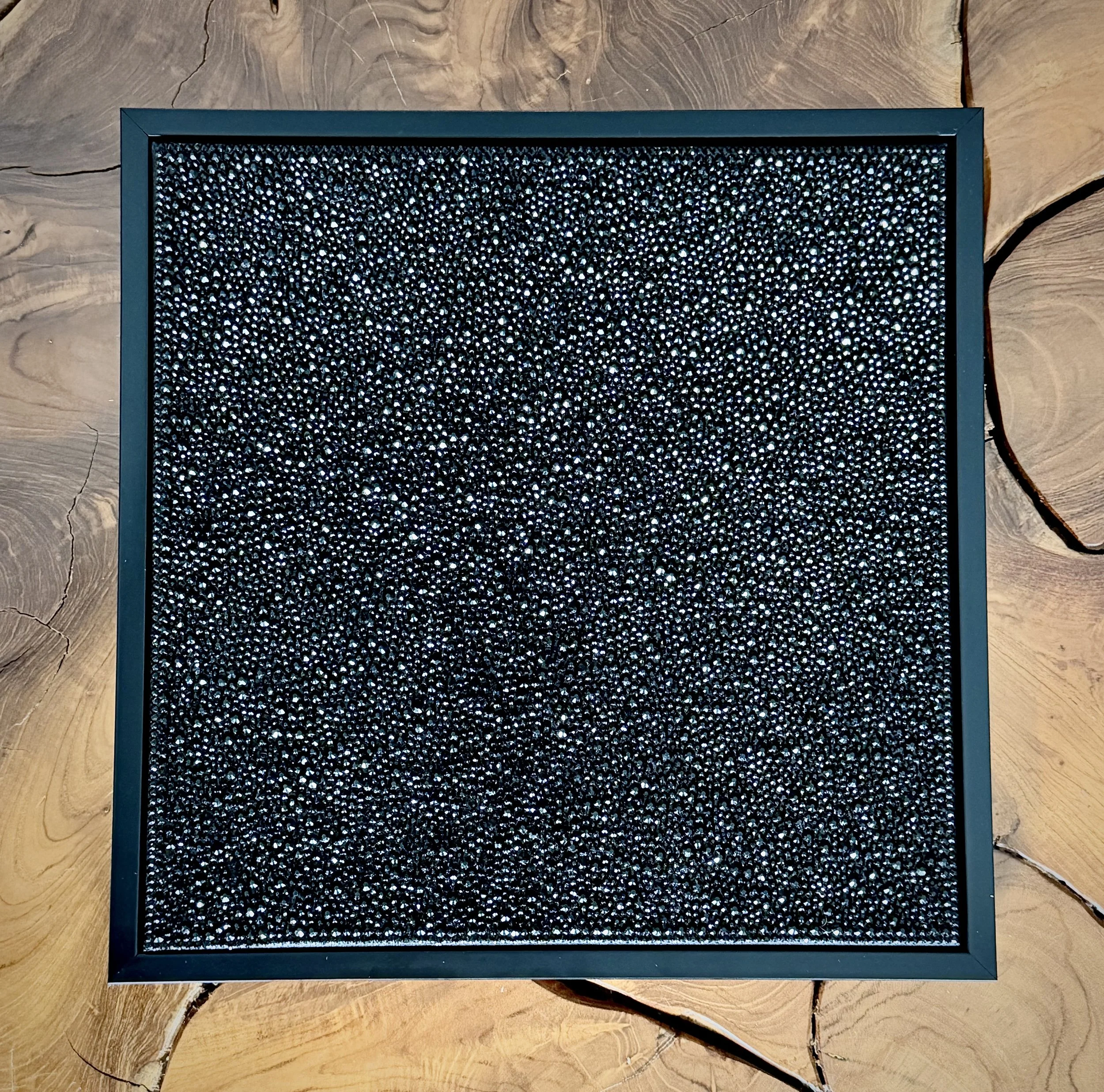 Pressure (2025) displayed on a modern wood coffee table. The 12x12 in. black panel’s dark texture contrasts with the natural grain.