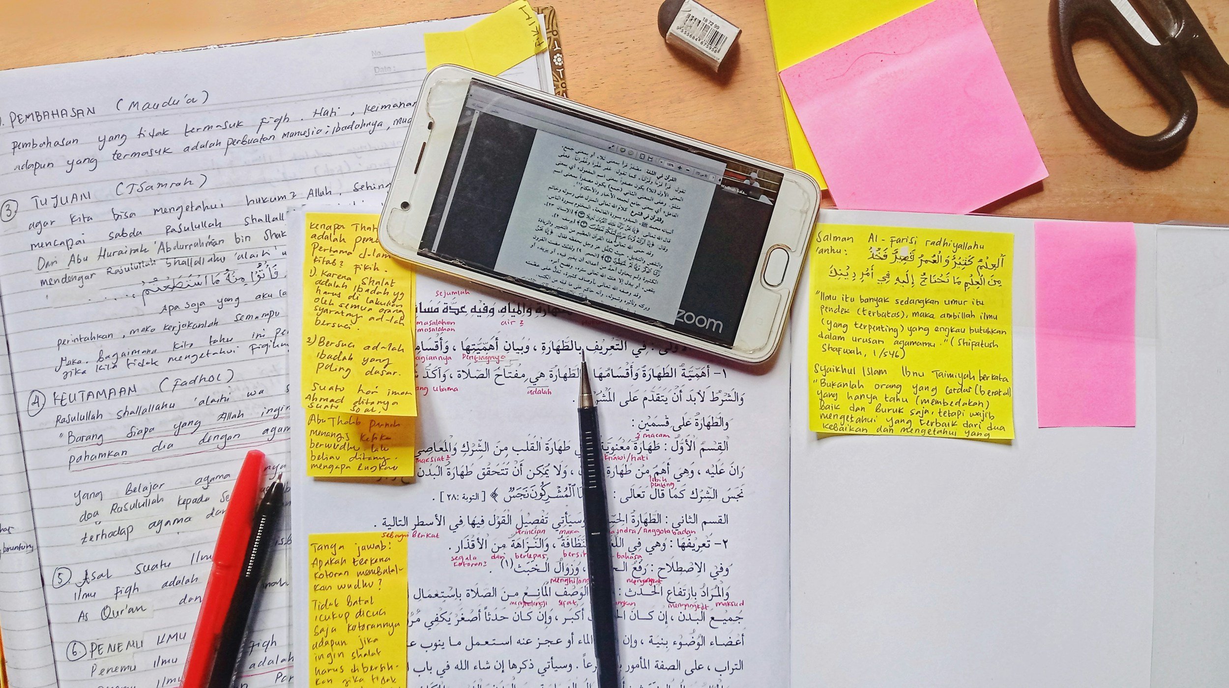 Study workspace with handwritten notes, post-it notes, a smartphone displaying text on an app, a pen, a red and black marker, scissors, a small eraser, and yellow, pink, and beige sticky notes on a wooden desk.