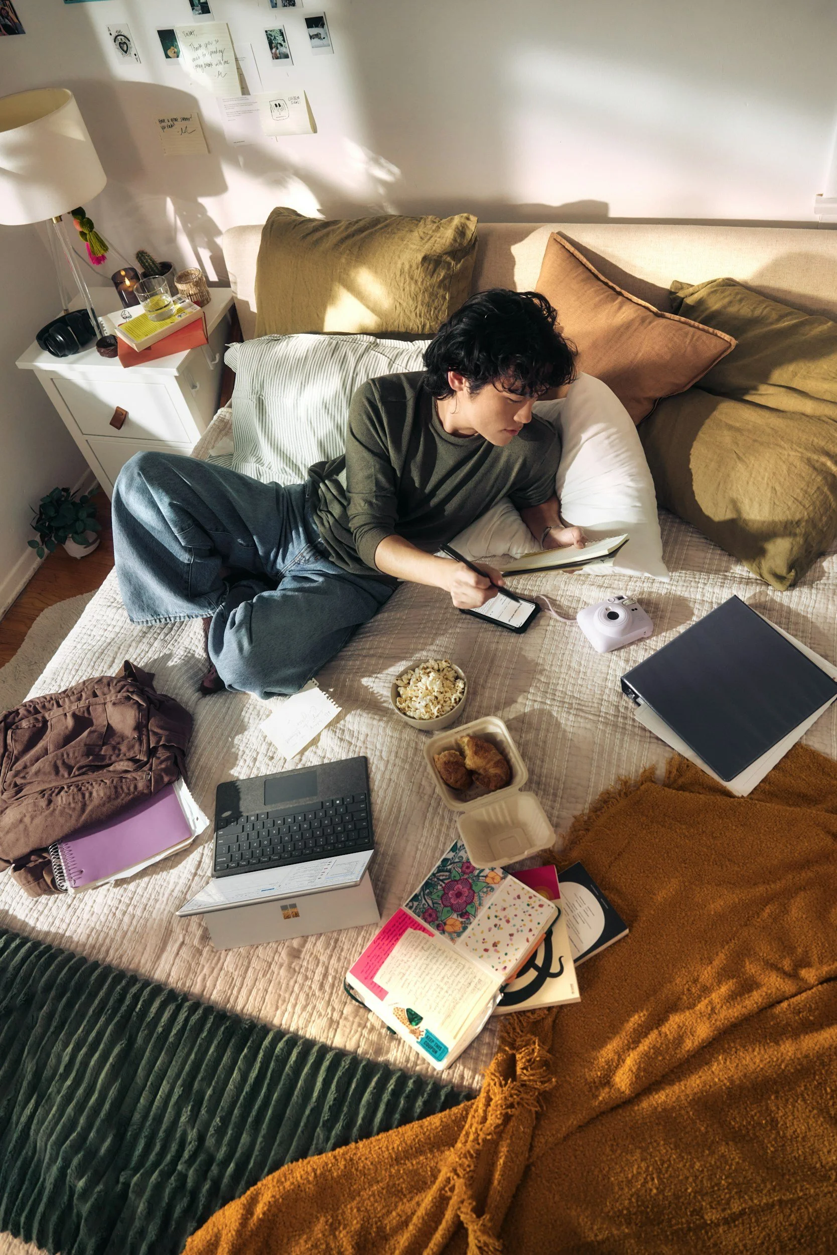 A person lying on a bed, reading a notebook. The bed has multiple pillows and is surrounded by various items including books, a bowl of popcorn, a container of fried chicken, a tablet, a phone, a camera, a small desk calendar, and a notebook. The room has a white wall with photographs and notes, a nightstand with items, and a green blanket and an orange blanket on the foot of the bed.