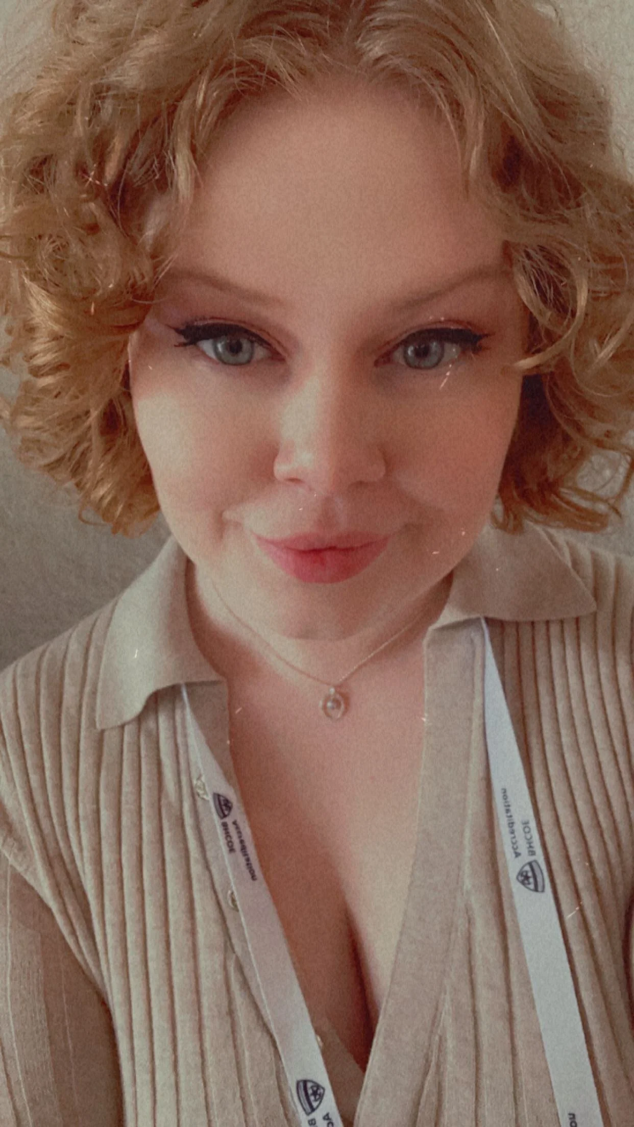 A woman with short curly red hair and blue eyes smiling at the camera, wearing a beige striped blazer, a white shirt, and a small necklace.