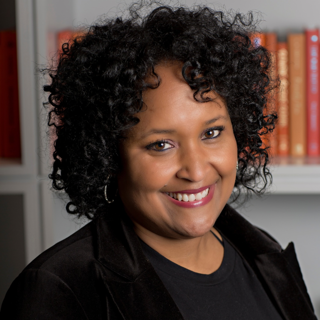 A woman with curly black hair smiling at the camera, wearing earrings and a black outfit, in a room with a bookshelf in the background.
