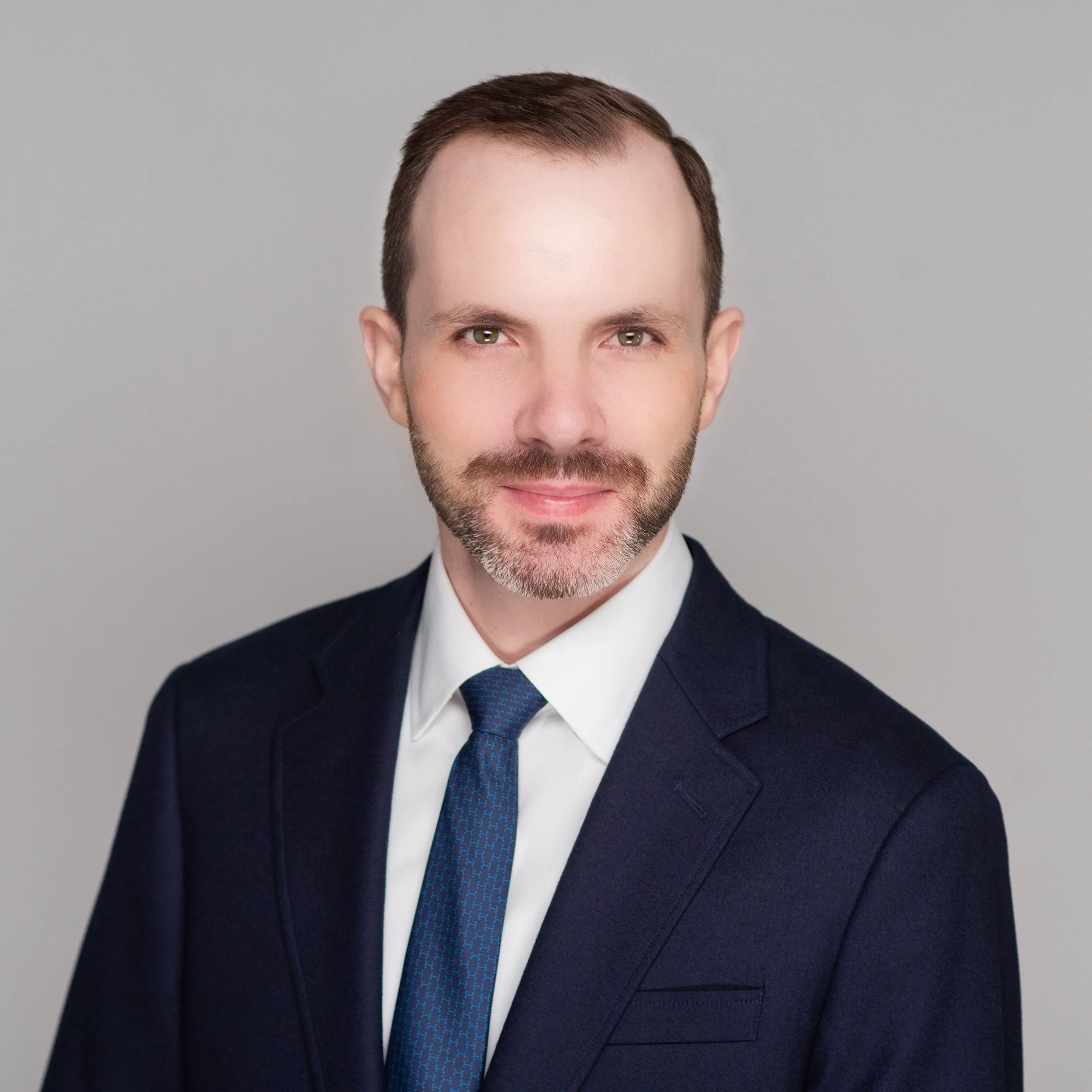 Professional headshot of a man with short brown hair, a trimmed beard, wearing a dark navy suit, white shirt, and a blue tie with subtle pattern, against a gray background.