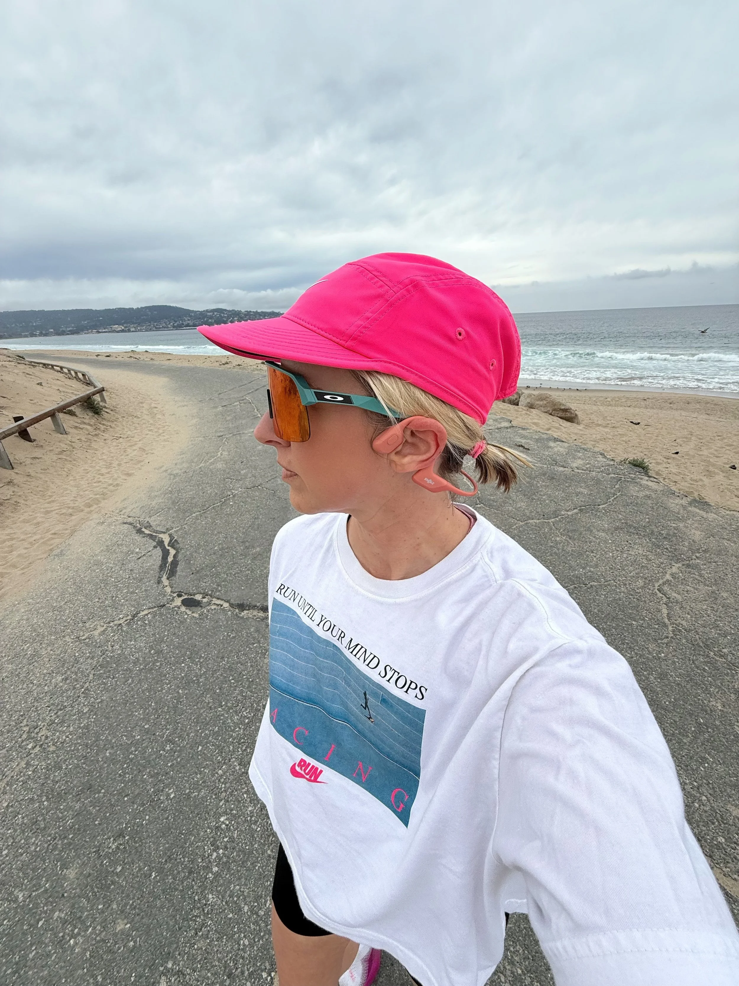 Side profile of a woman running along a beach path under cloudy sky, wearing a pink cap, sunglasses, a white T-shirt, and black shorts.