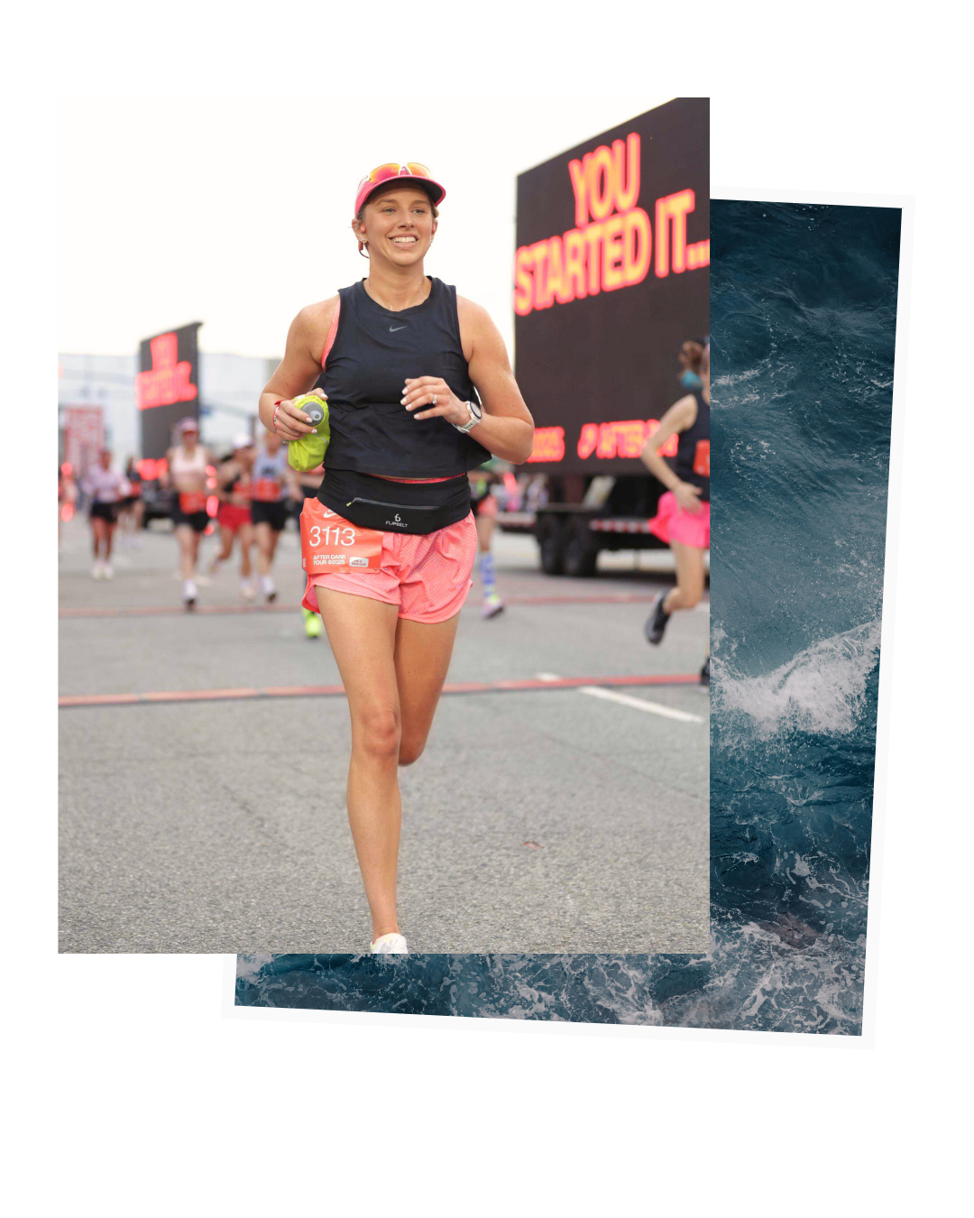 Female runner participating in a marathon, smiling and holding a water bottle, wearing a pink cap, black tank top, and pink shorts with the race bib 3113, surrounded by other runners and a large electronic billboard in the background.
