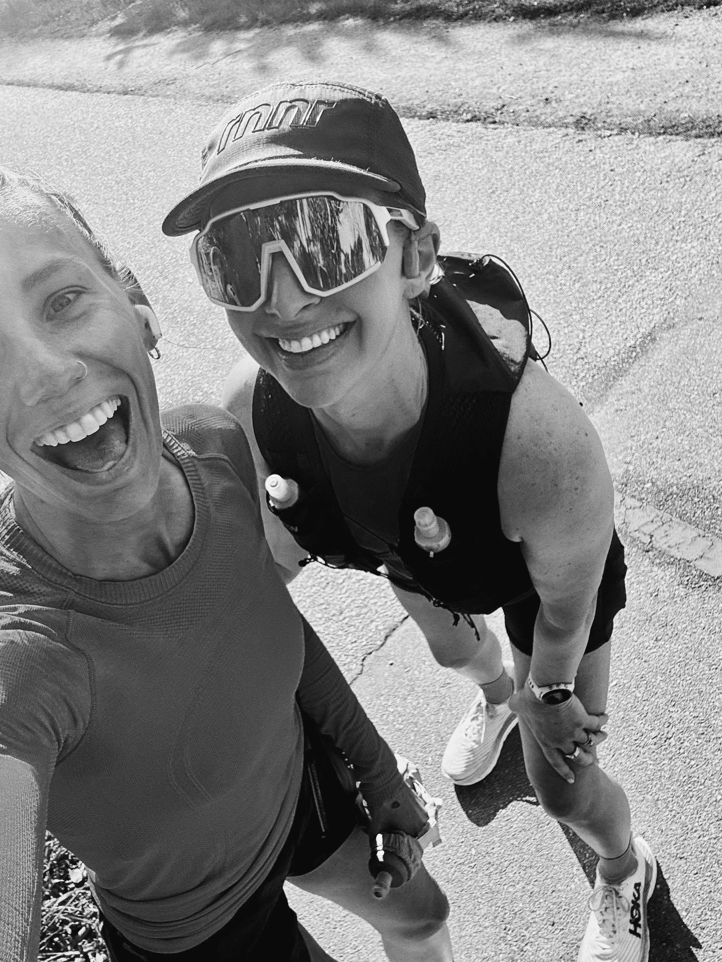 Two friends smiling, taking a selfie outdoors on a sunny day. One person is wearing a cap, large sunglasses, and a sleeveless athletic top, while the other has earrings and an athletic top. They are standing on a paved surface with some grass visible in the background.
