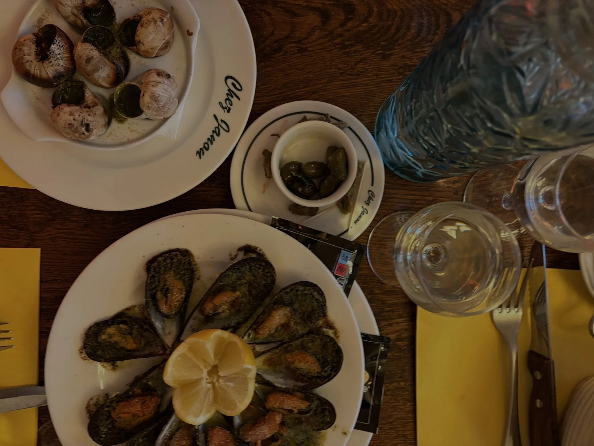 A table with plates of cooked mussels garnished with lemon wedges in the center, a small dish of olives, a glass of water, and two empty wine glasses set with yellow napkins.