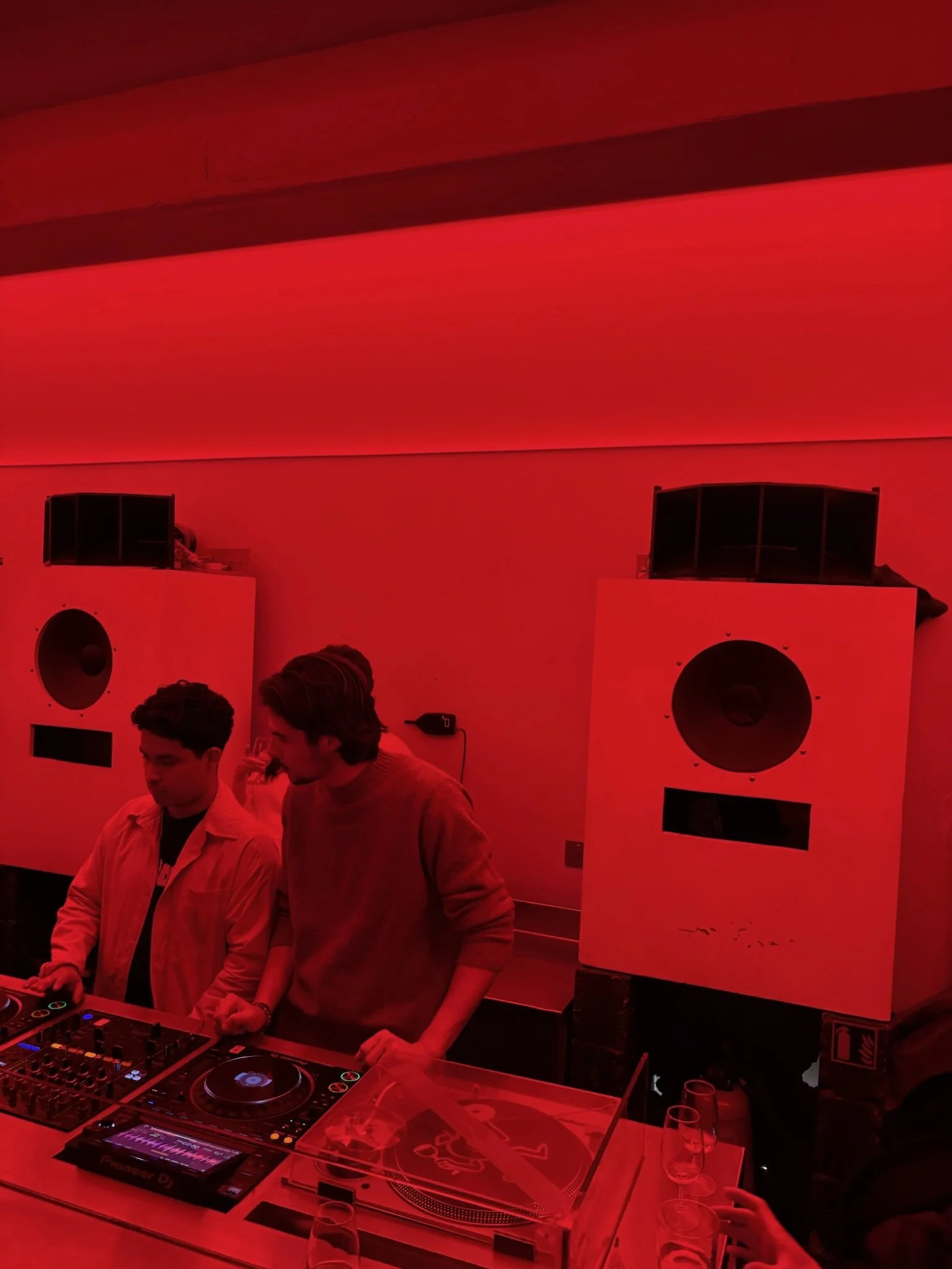 Two young men DJing at a party with red lighting, large speaker speakers behind them, DJ equipment, and glasses on the table in the foreground.