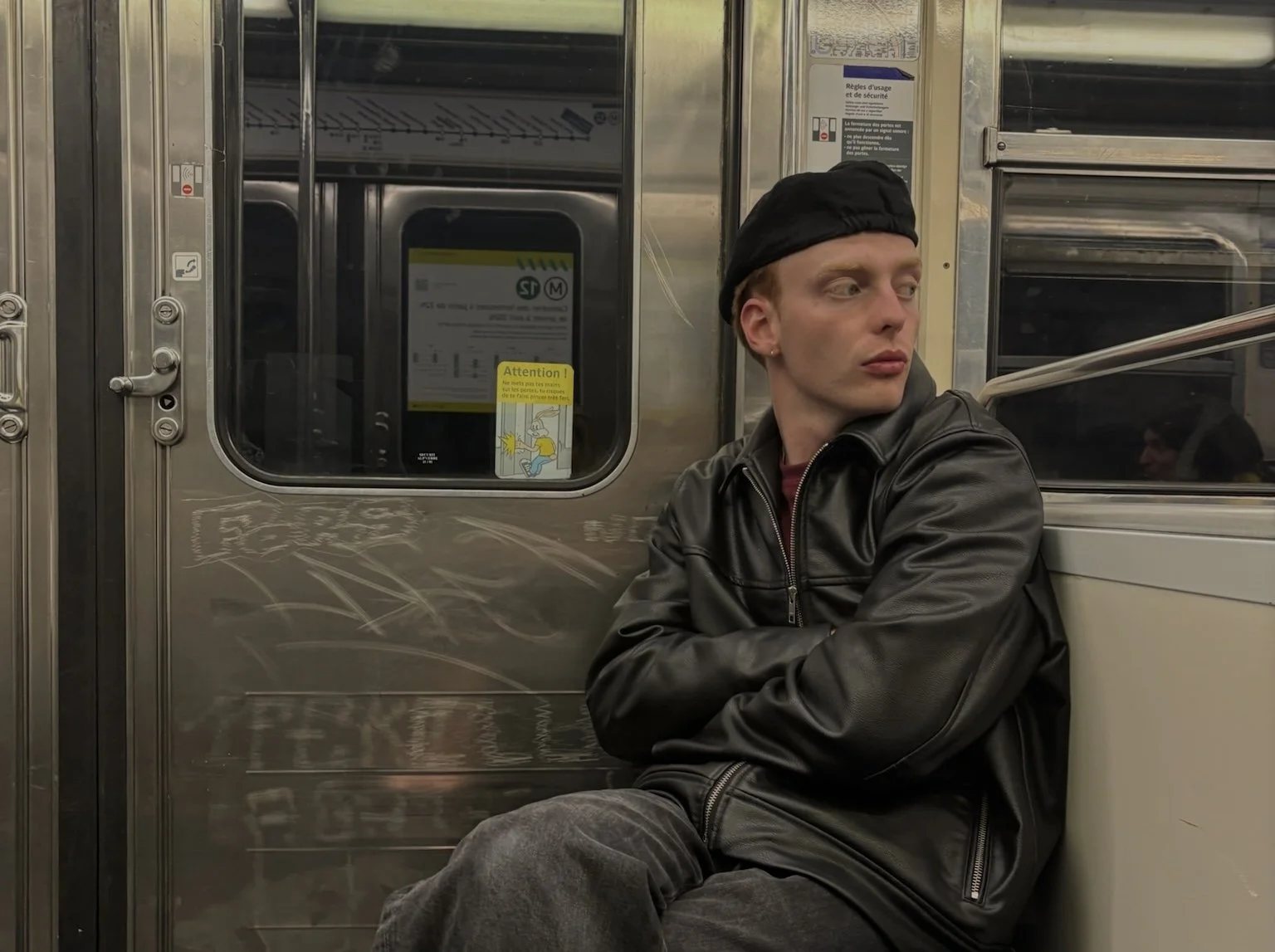 A young man with light skin, short hair, wearing a black leather jacket and a black beanie, sitting alone on a train with crossed arms, looking to his right.