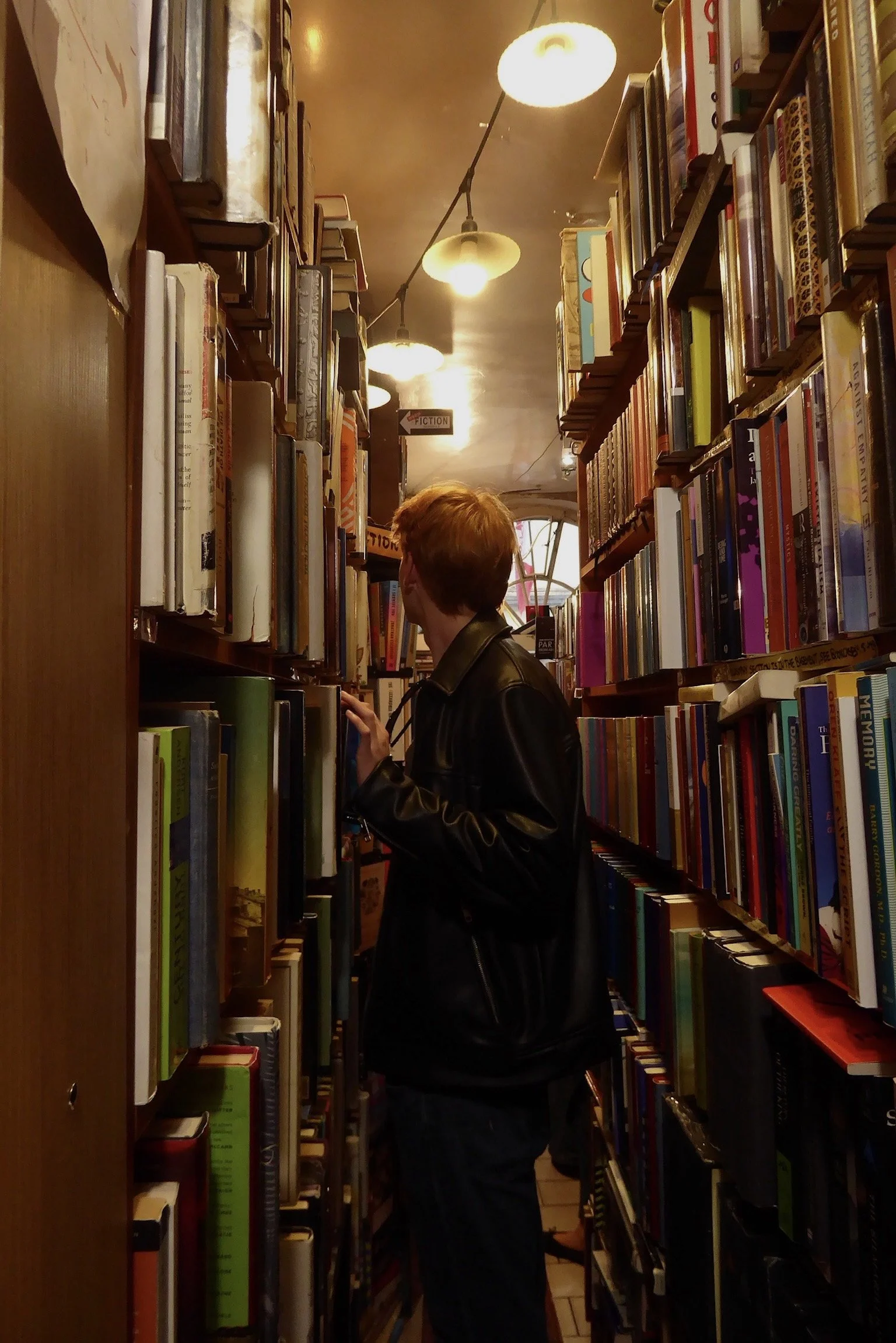 A person with red hair wearing a black leather jacket browsing books in a narrow bookstore aisle.