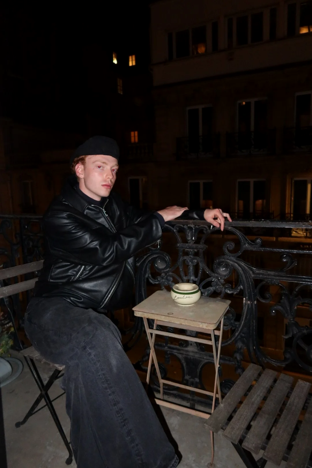 A young man in a black leather jacket and black beanie sitting on a balcony at night, with a small table and bowl in front of him, overlooking a building with lit windows.