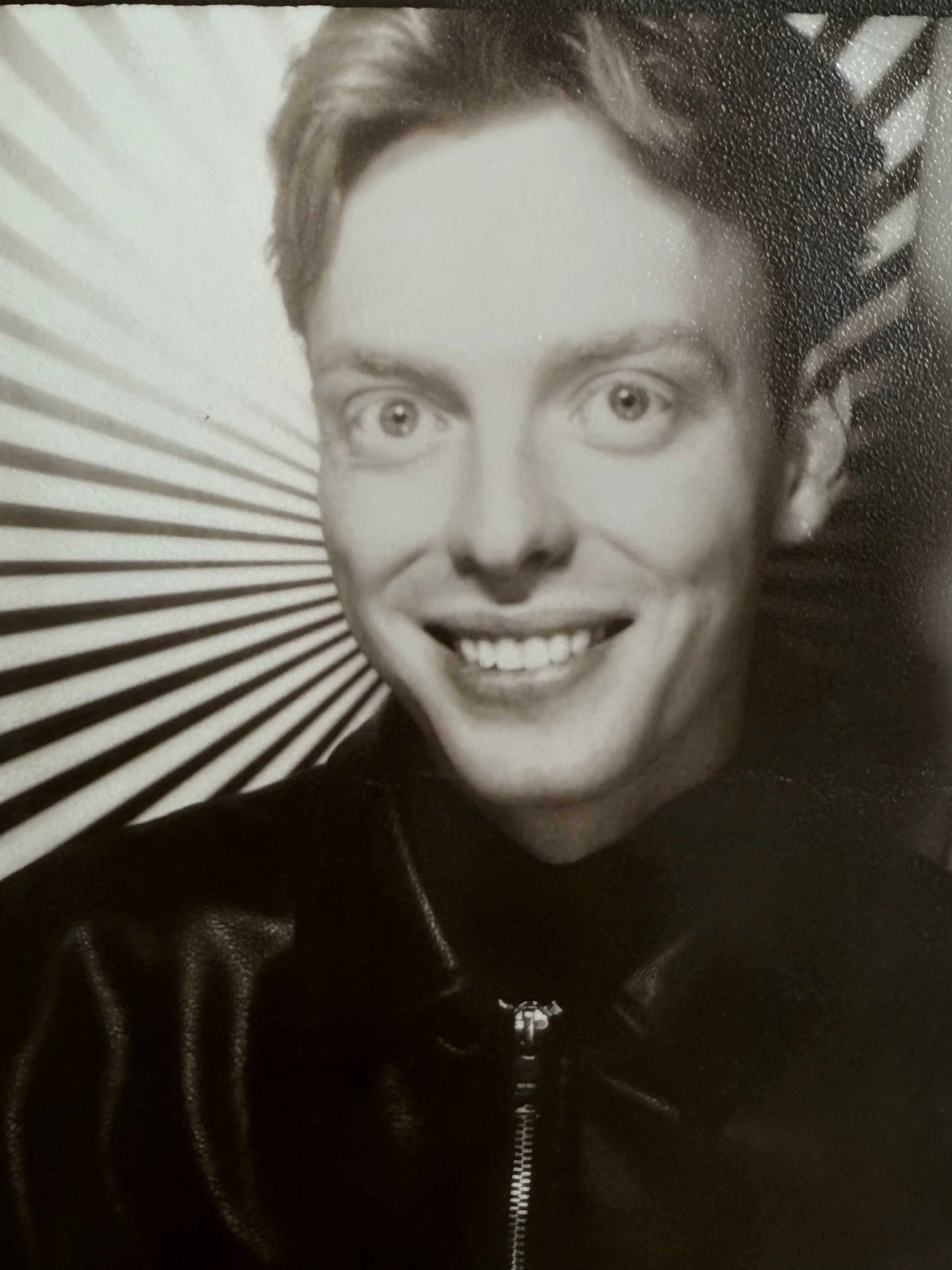 Black and white portrait of a smiling woman with short hair, wearing a leather jacket, and a striped background.