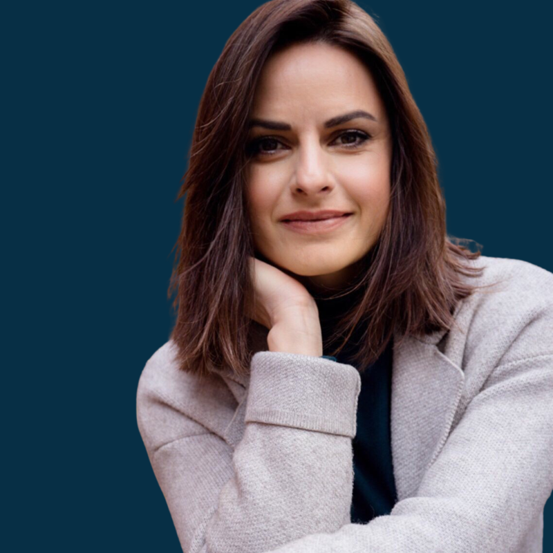 Portrait of a woman with shoulder-length brown hair, wearing a light gray blazer and smiling gently, with her right hand touching her neck, against a dark blue background.