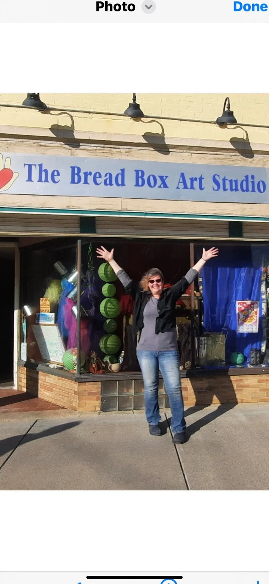 A woman standing outside a storefront called 'The Bread Box Art Studio' with her arms raised, smiling, in front of colorful decorations and artwork displayed in the window.