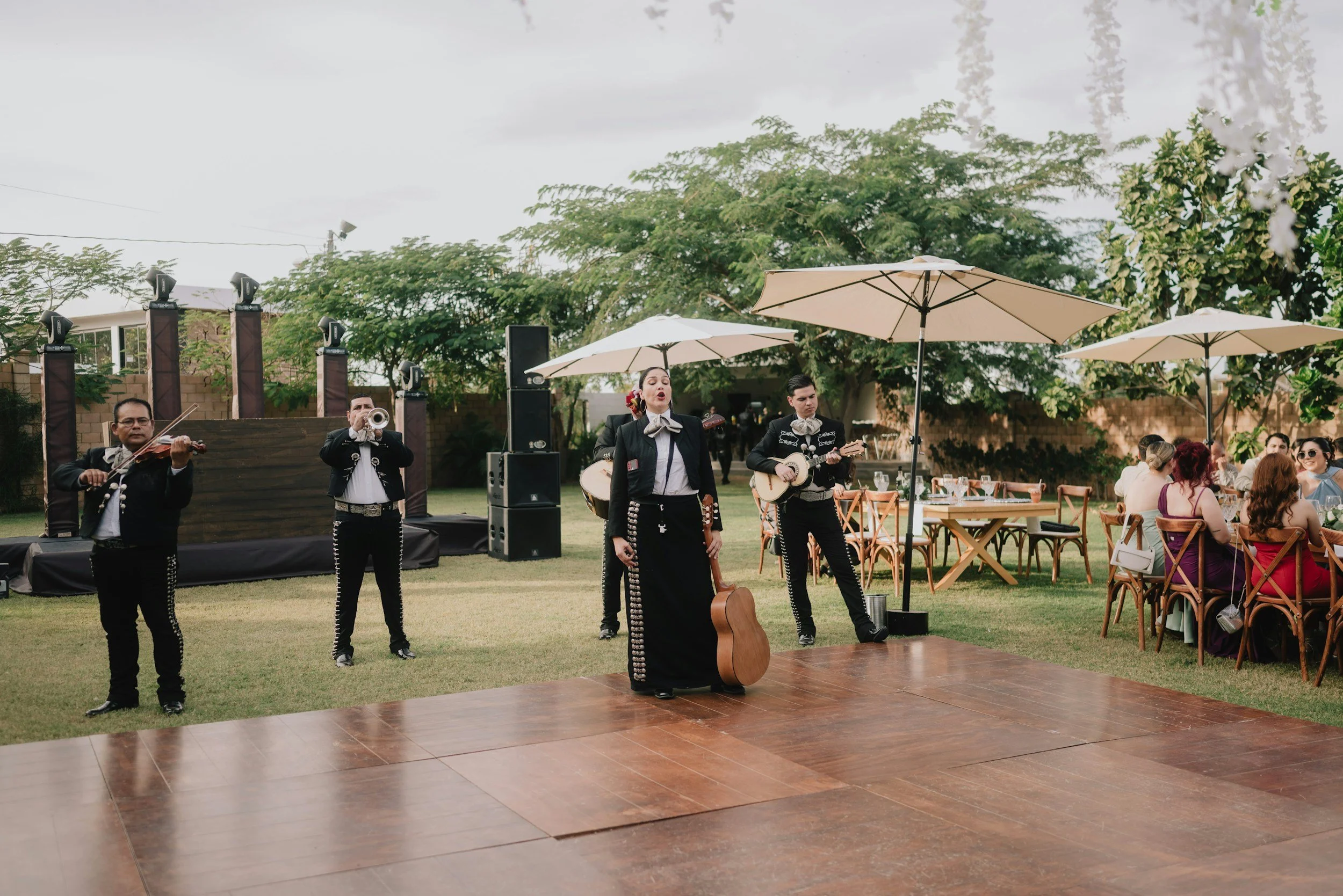 Grupo de músicos con trajes tradicionales mexicanos tocando instrumentos en un evento al aire libre con mesas, sillas y paraguas blancos, y muchas personas asistentes.