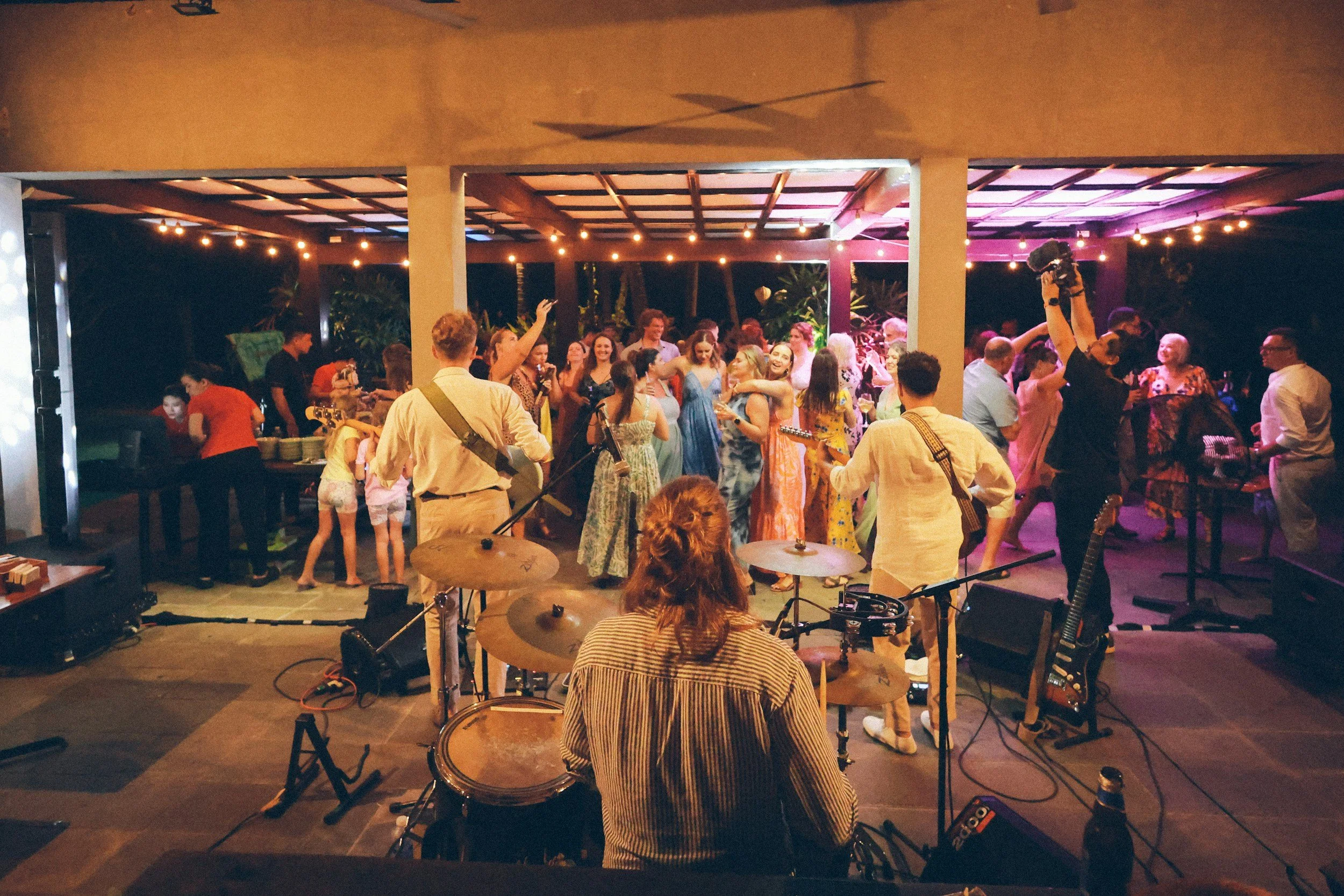 Una fiesta o concierto con muchas personas bailando y disfrutando en un espacio cubierto decorado con luces. En el escenario, una banda tocando instrumentos, visible por los instrumentos de batería y guitarras, con una multitud de personas sonrientes y bailando en segundo plano.