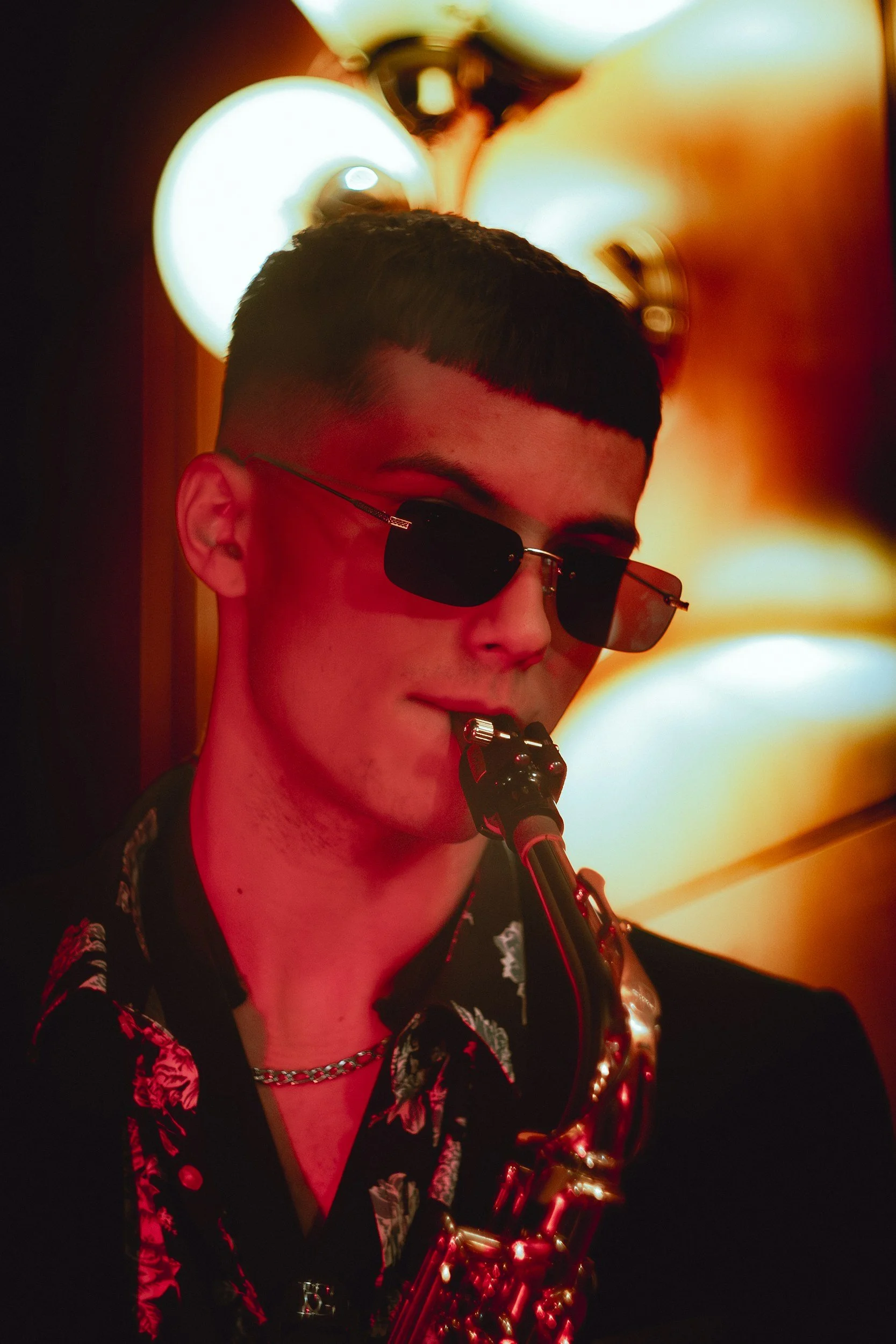 Un joven tocando un saxofón en un entorno con iluminación cálida y ambiente artístico, lleva gafas de sol negras y una camisa negra con patrones de flores coloridas.