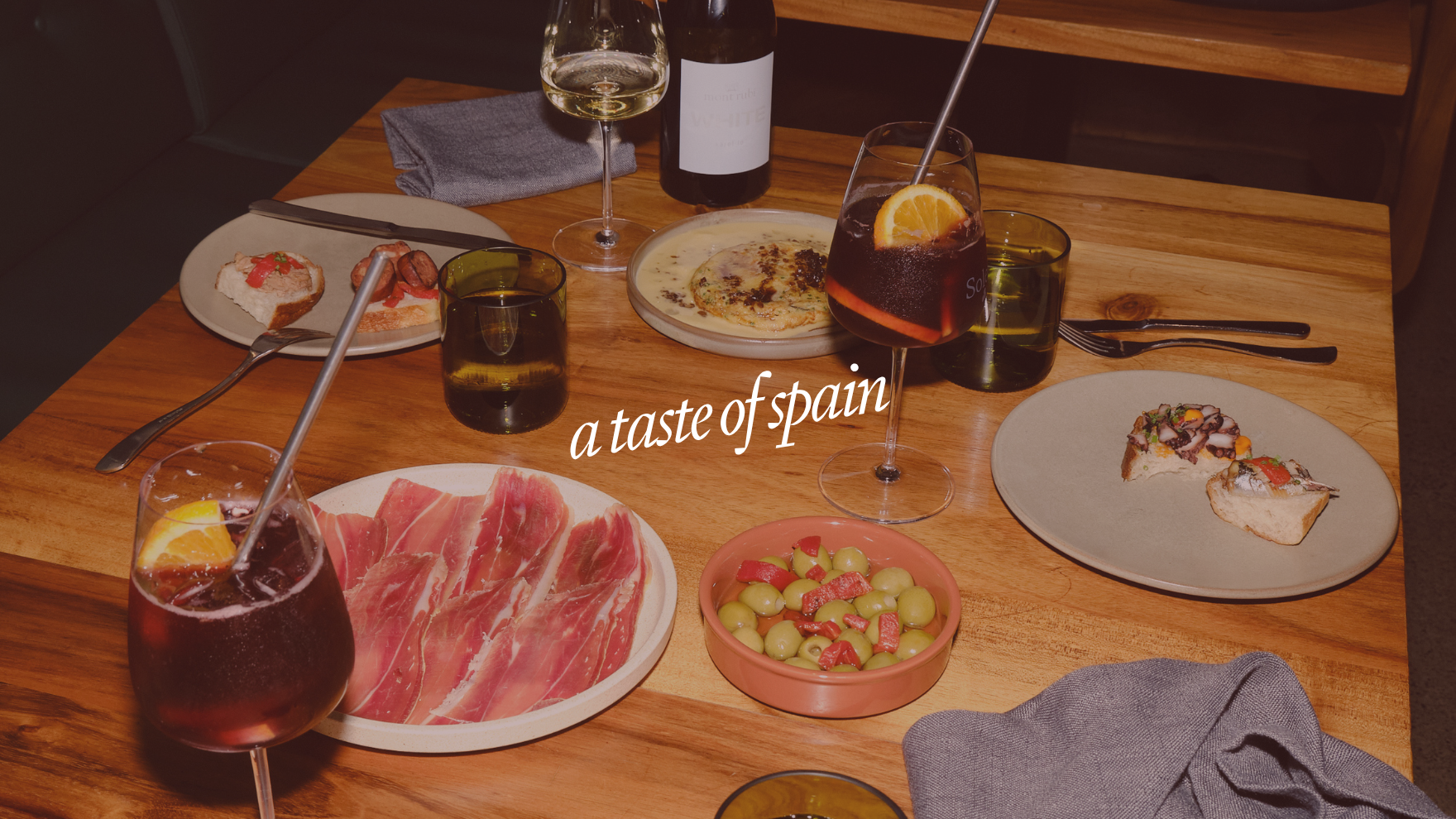 A wooden table set with glasses of red and white wine, plates of Spanish tapas including sliced cured ham, assorted canapés, a bowl of olives and chopped peppers, a bottle of white wine, a bowl of a creamy dish, and a glass of sangria with orange slices. The words "a taste of spain" are overlaid on the image.