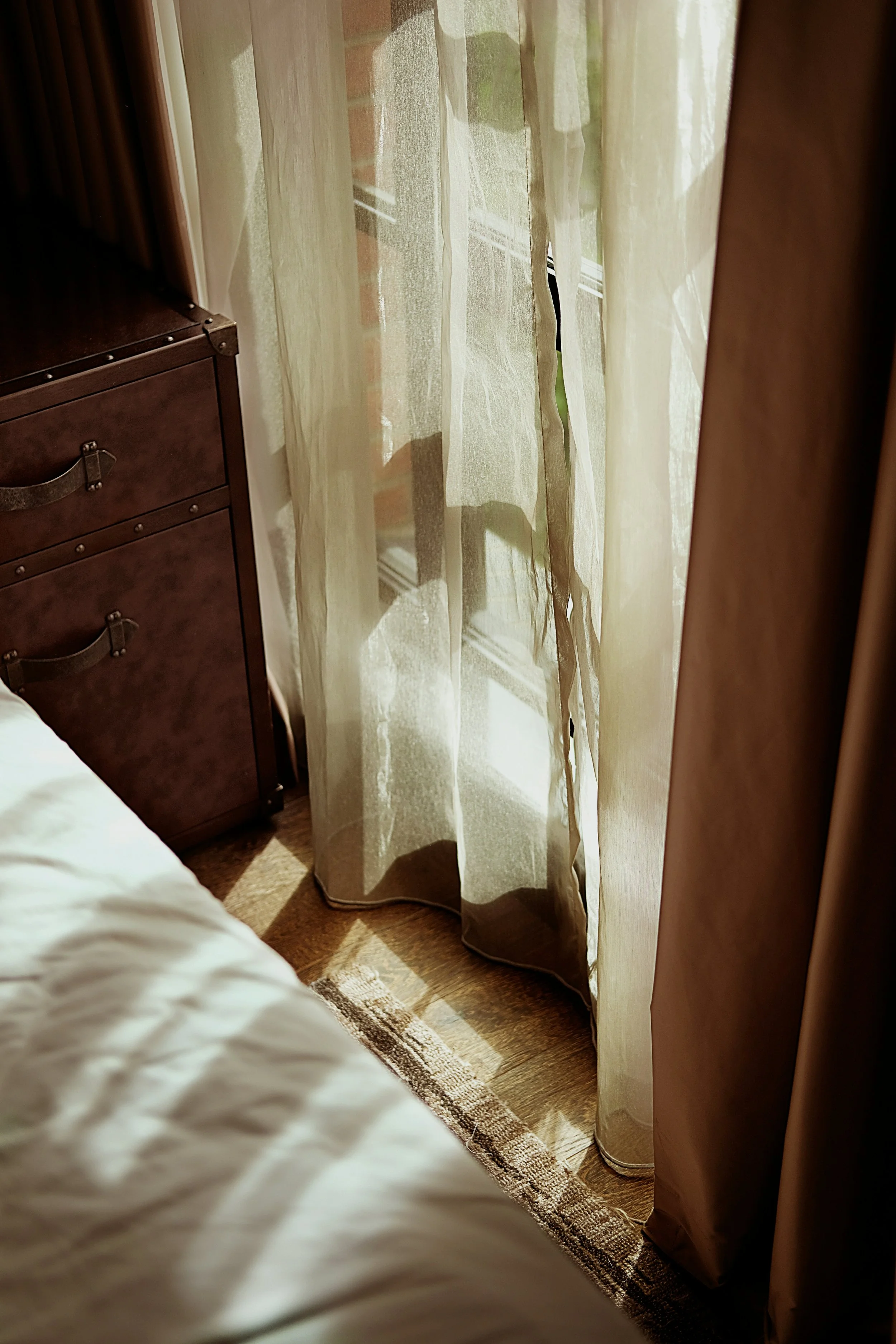 Ein Schlafzimmer mit Bett, einem Nachtschrank, Vorhängen, die Sonnenlicht hereinlassen, samt Teppich auf Holzboden.