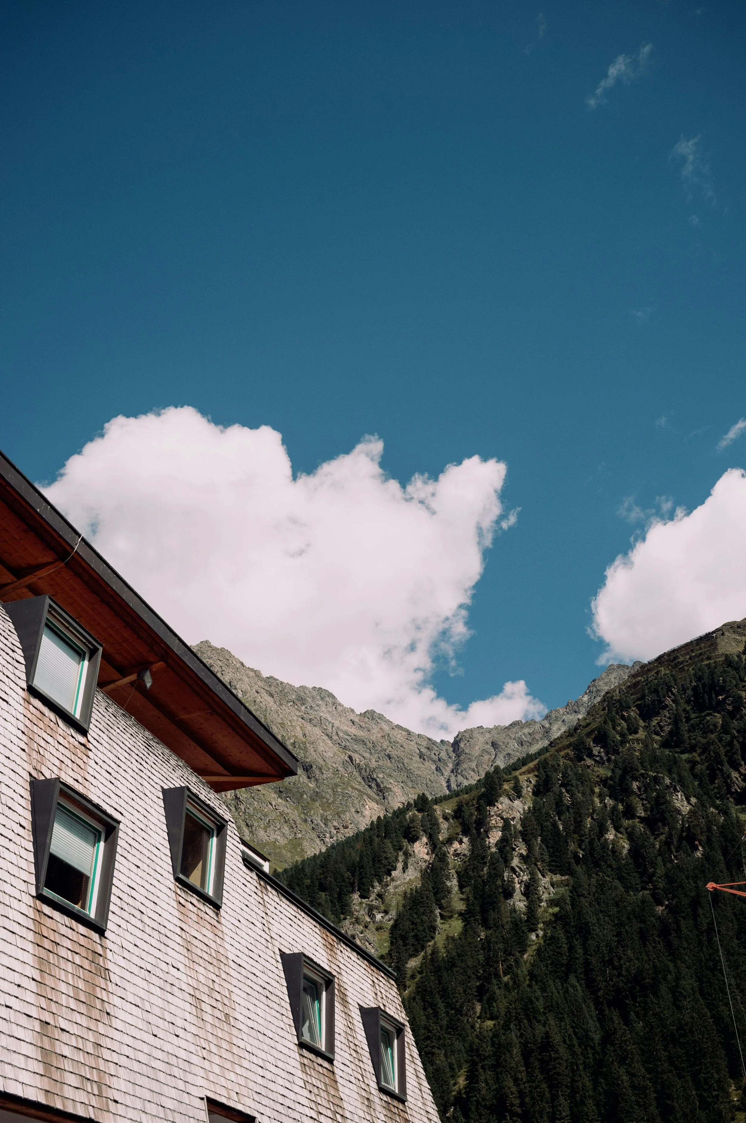 Ein Haus mit Schindel-Fassade und mehreren Fenstern, im Hintergrund Berge und ein blauer Himmel mit Wolken.