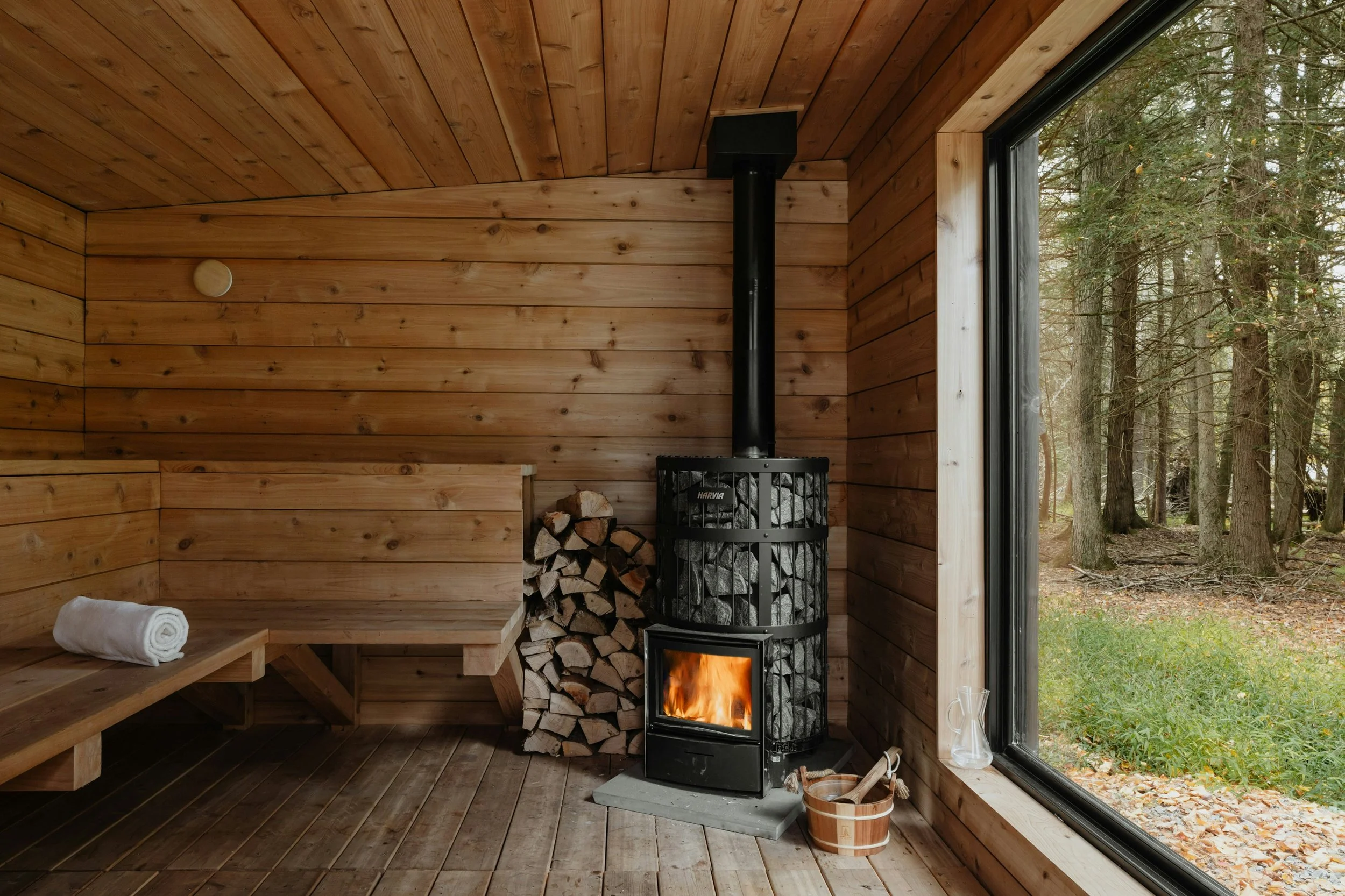 Innenraum einer Sauna mit Ofen, einem Stapel mit Feuerholz, einer Bank mit Handtuch und einem großen Fenster, das einen Wald draußen zeigt.