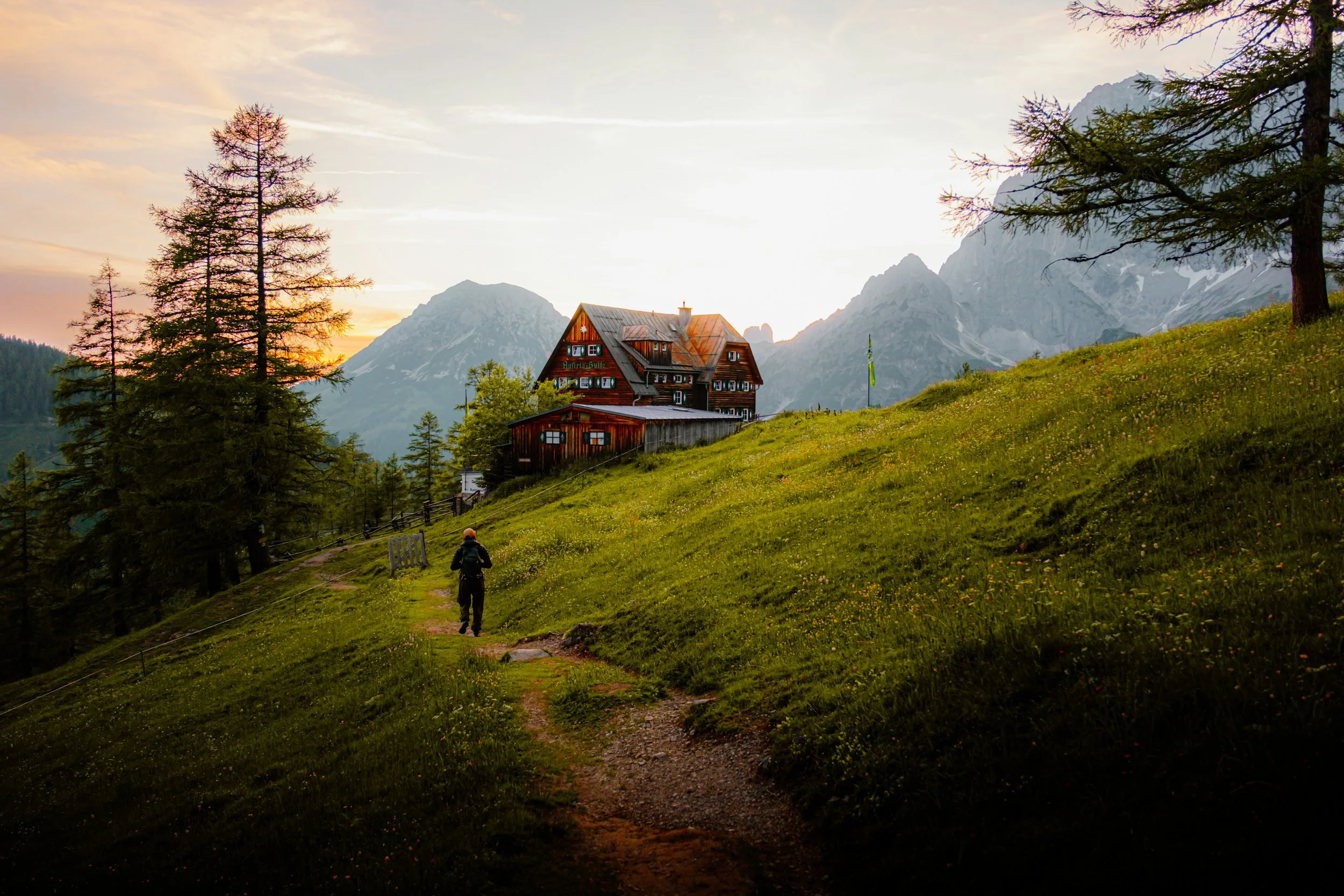 Berglandschaft mit einem Pfad, einem Wanderer, einer Holzhütte, grünen Wiesen, Bäumen und Bergen im Hintergrund bei Sonnenuntergang.