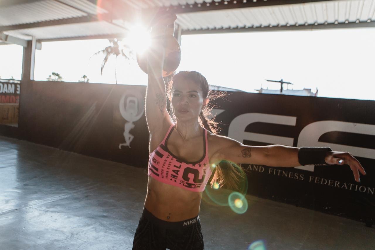 A woman in athletic wear performing a workout inside a gym with sunlight streaming in, holding a kettlebell overhead.