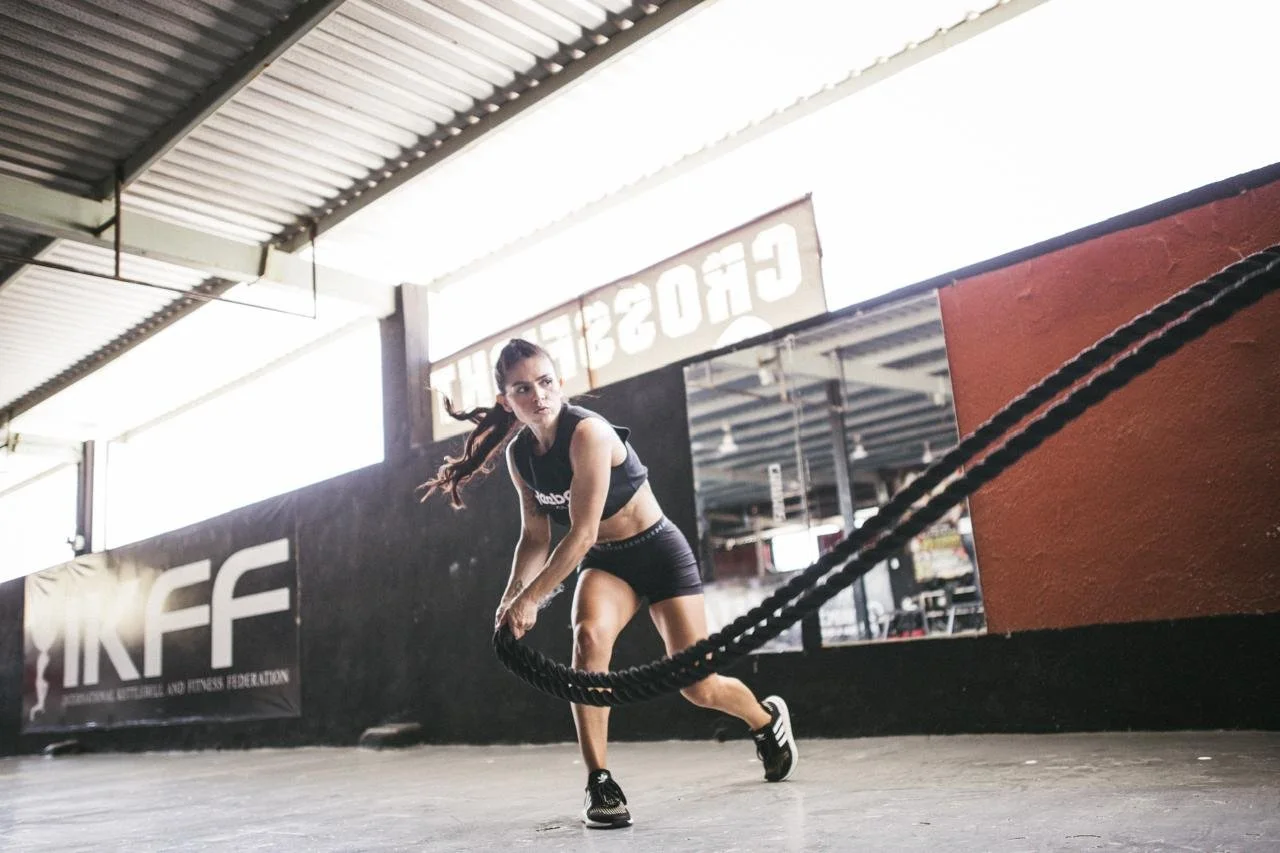 A woman in workout attire is training with battle ropes inside a gym.
