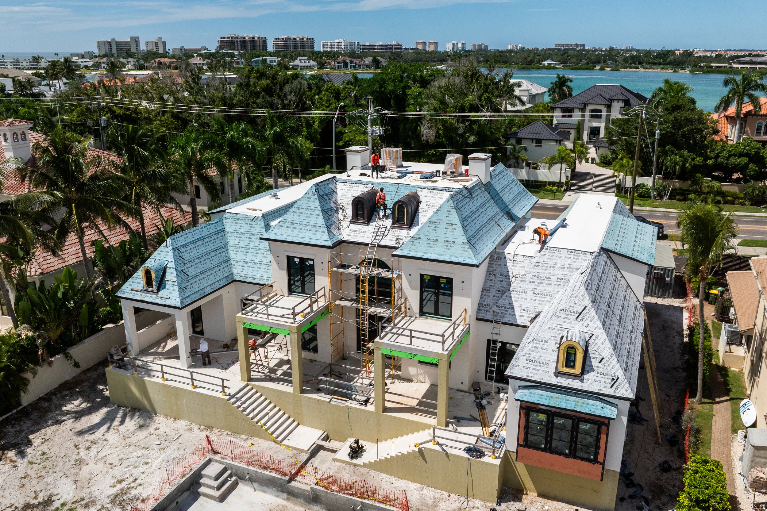 A large house under construction with workers on the roof, scaffolding, and a partially finished front porch. The roof has blue underlayment, and the surrounding area features neighboring houses, palm trees, and a body of water in the background.