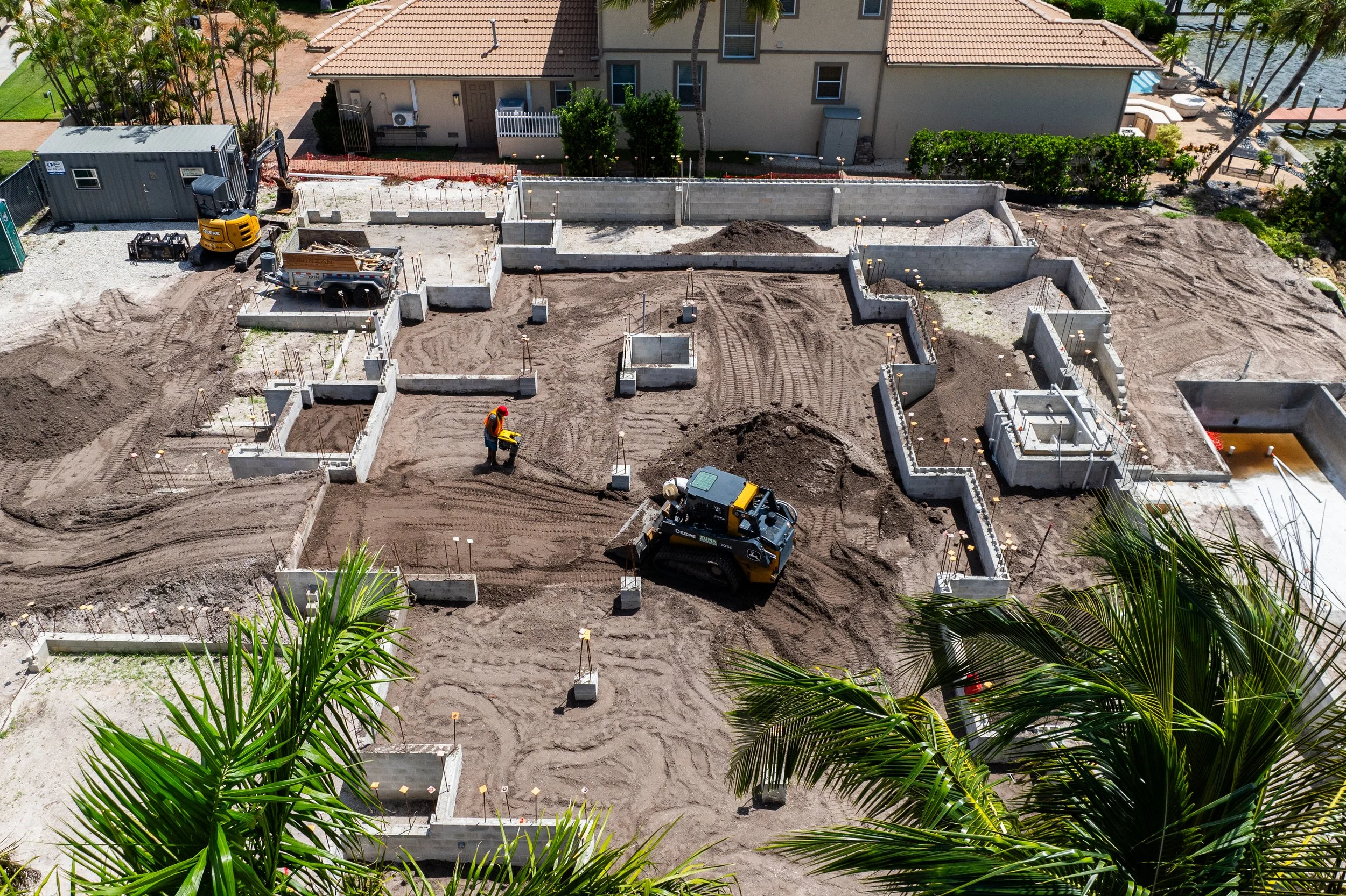 Construction site with foundation being laid, workers and machinery, surrounding residential buildings, lush greenery, and palm trees.