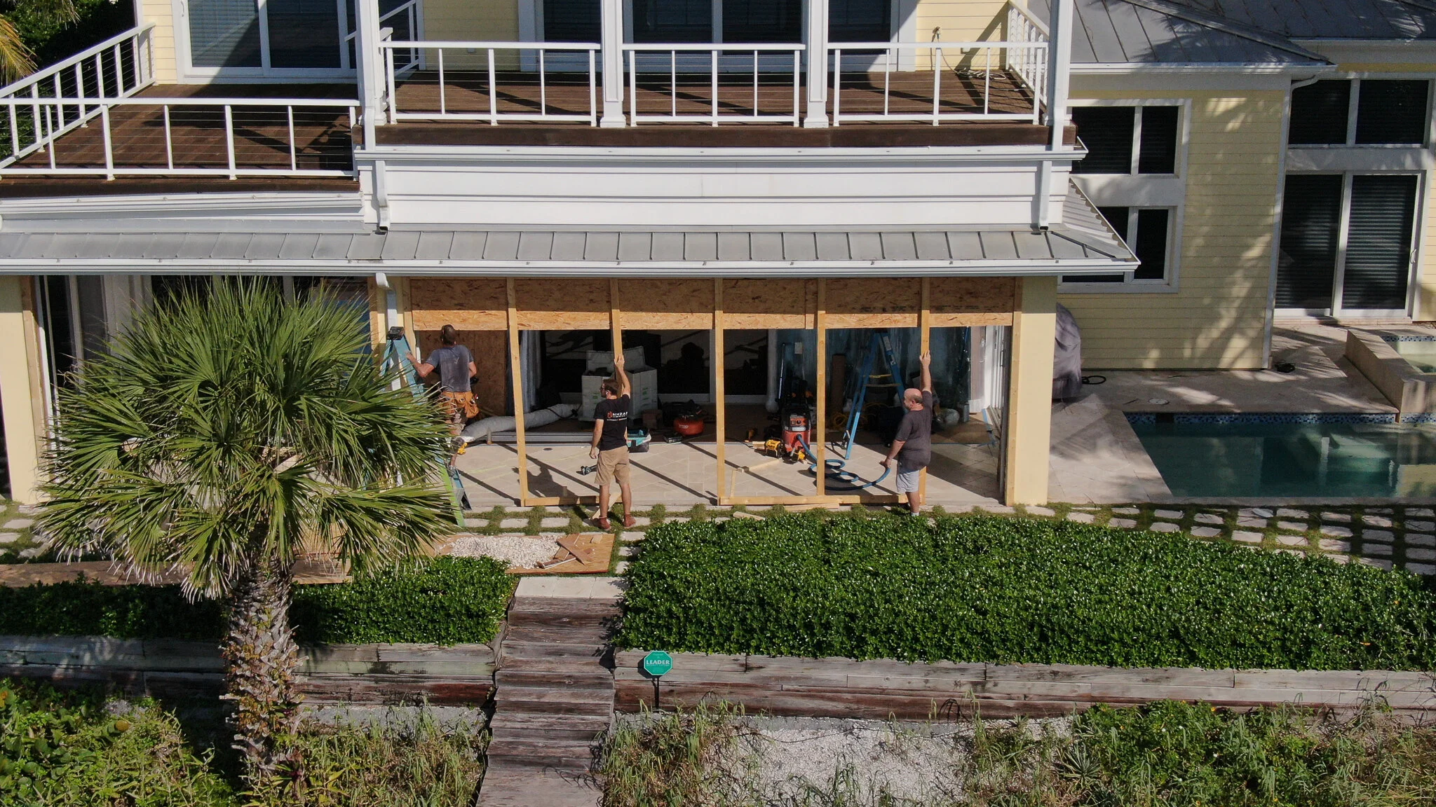 A group of people working on constructing a screened porch on a yellow house with sliding glass doors, with a pool in the backyard and a palm tree in the foreground.