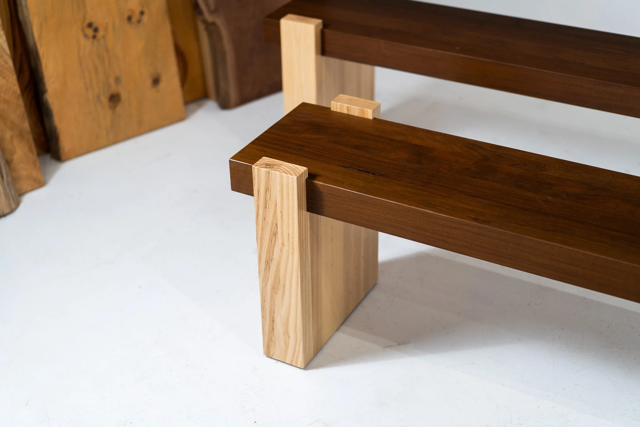 Close-up of a wooden bench with a dark brown seat and a light-colored wooden leg, on a white floor with wood planks in the background.