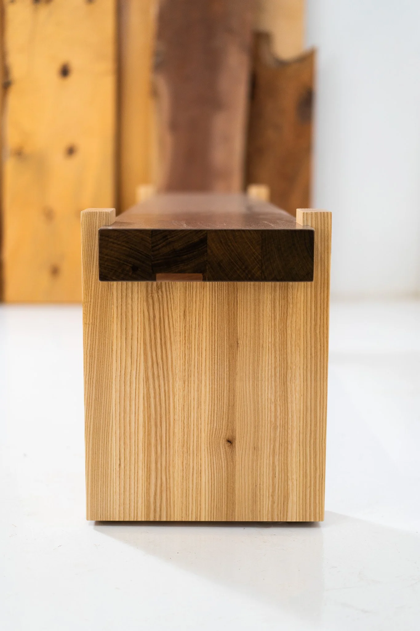 Close-up of a wooden bench with a natural finish, showing the end and seat surface, with a blurred background of wooden planks.