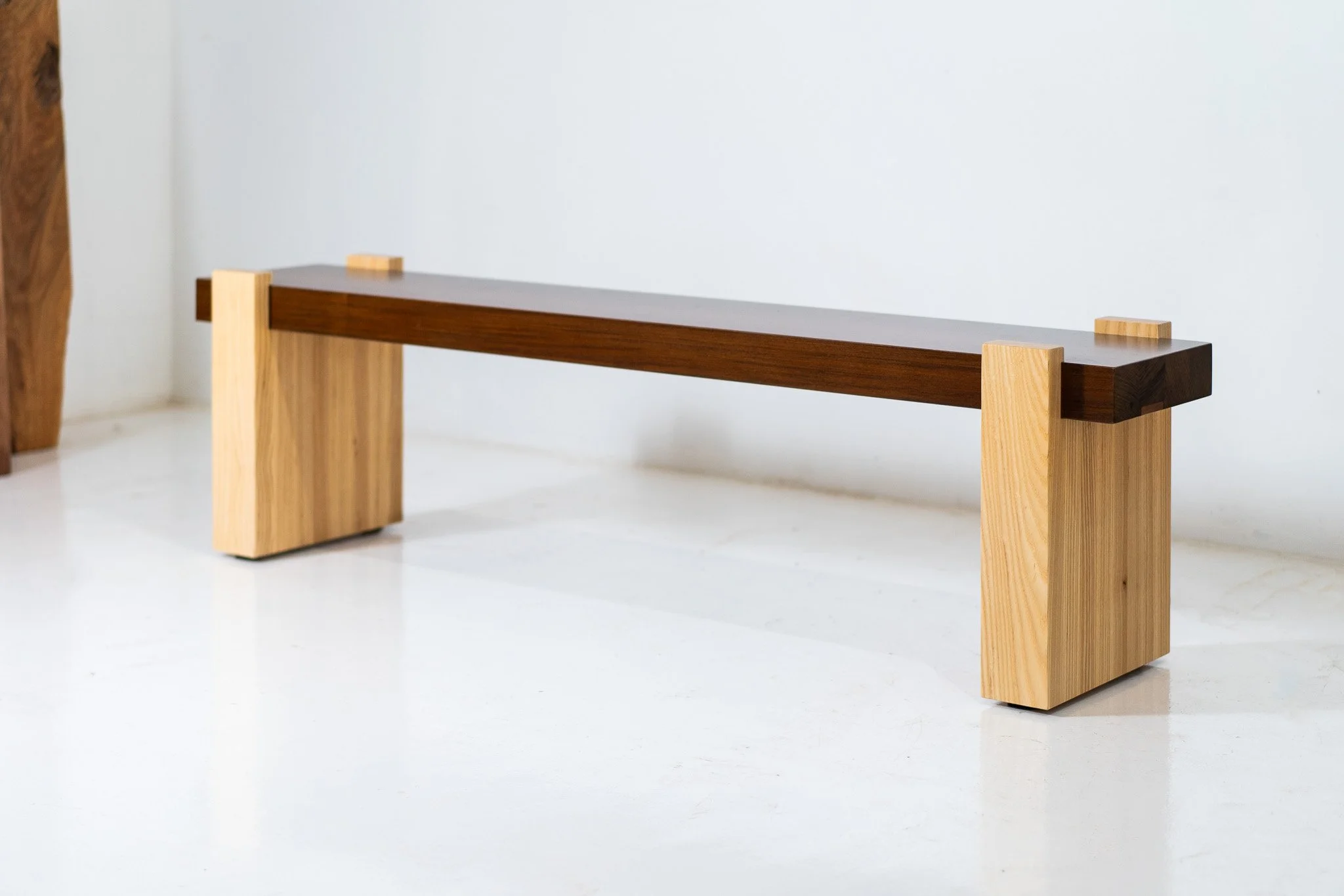 Modern wooden bench with a long dark brown seat and lighter wood legs against a white wall.