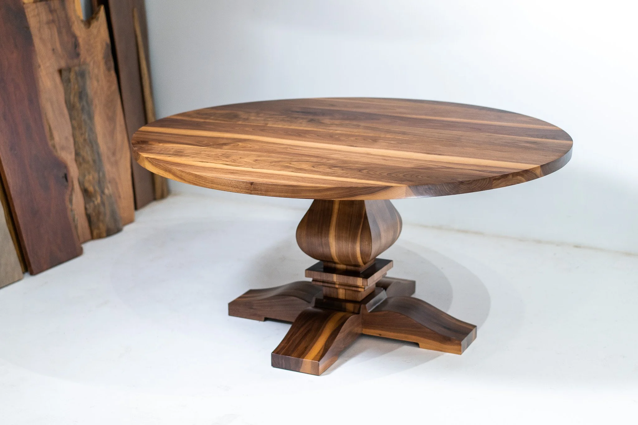 A round wooden table with a pedestal base on a white surface, with wood planks leaning against a wall in the background.