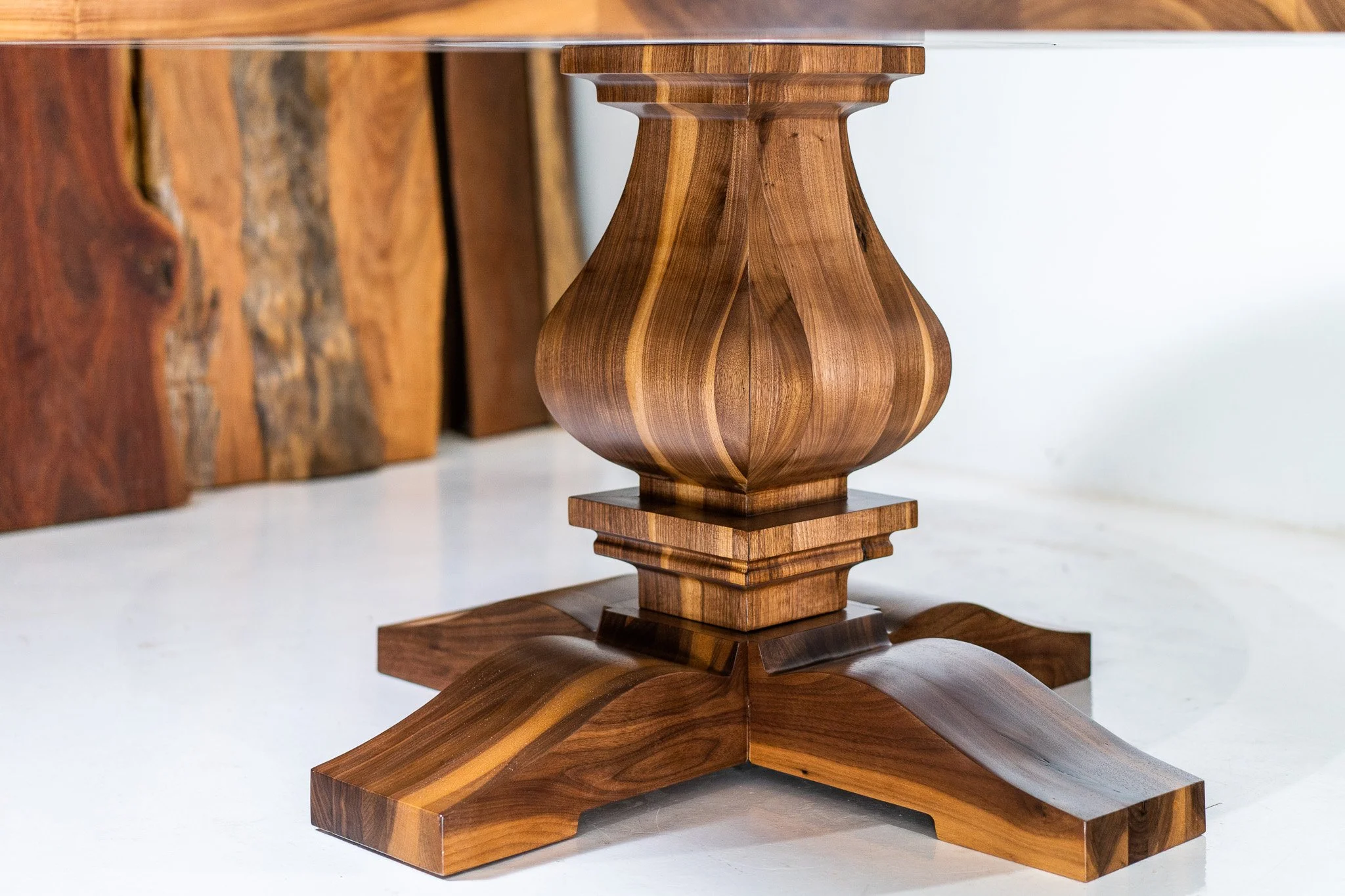 Close-up of a wooden table base with a curvaceous, bulbous shape, sitting on a white surface with a background of other wooden furniture pieces.