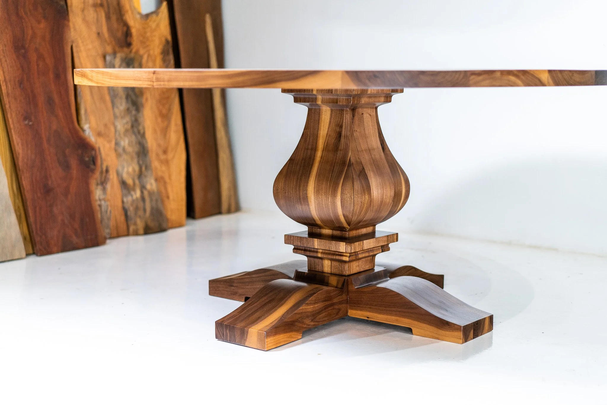 A wooden table with a rounded, decorative central support and four wide, curved legs, placed on a white floor with wooden planks leaning against the wall in the background.