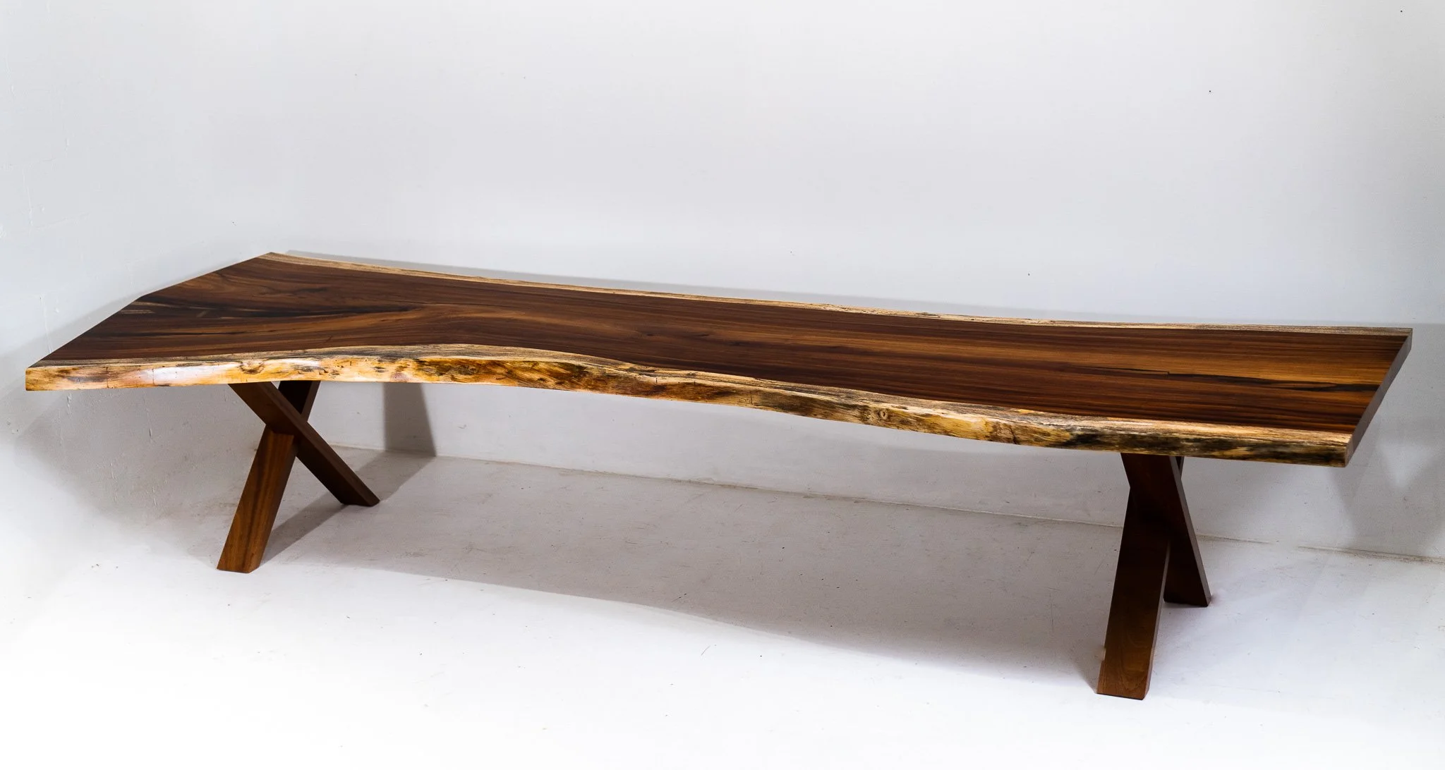 A modern wooden bench with a natural live edge design, featuring a polished surface and two angular legs, set against a plain white wall.