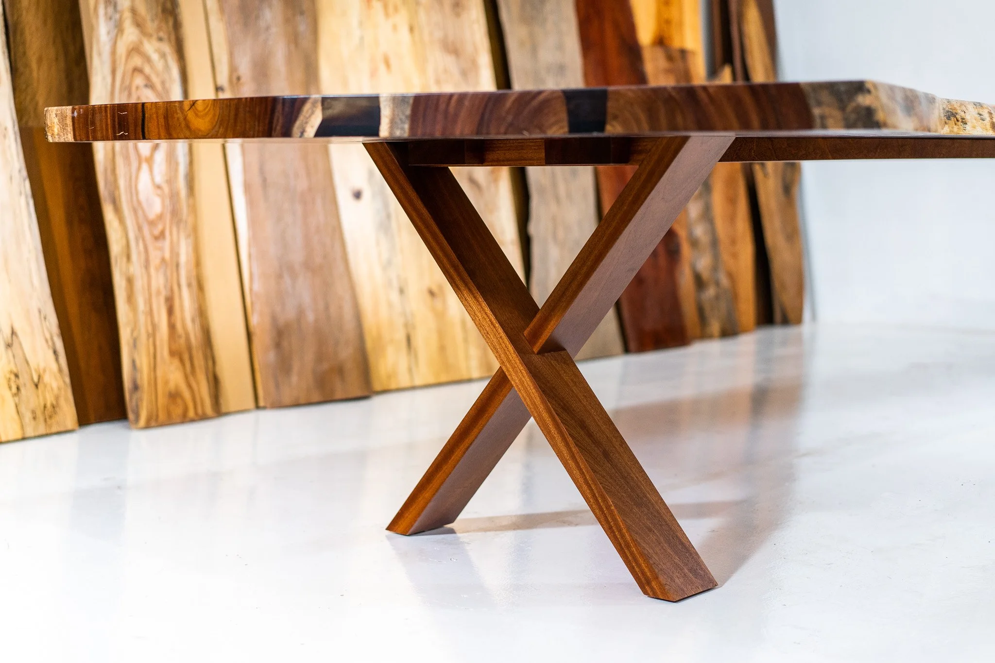 Close-up view of a wooden table with a natural finish, showing the edge and the unique X-shaped leg design. Background features various wooden planks leaning against a wall.