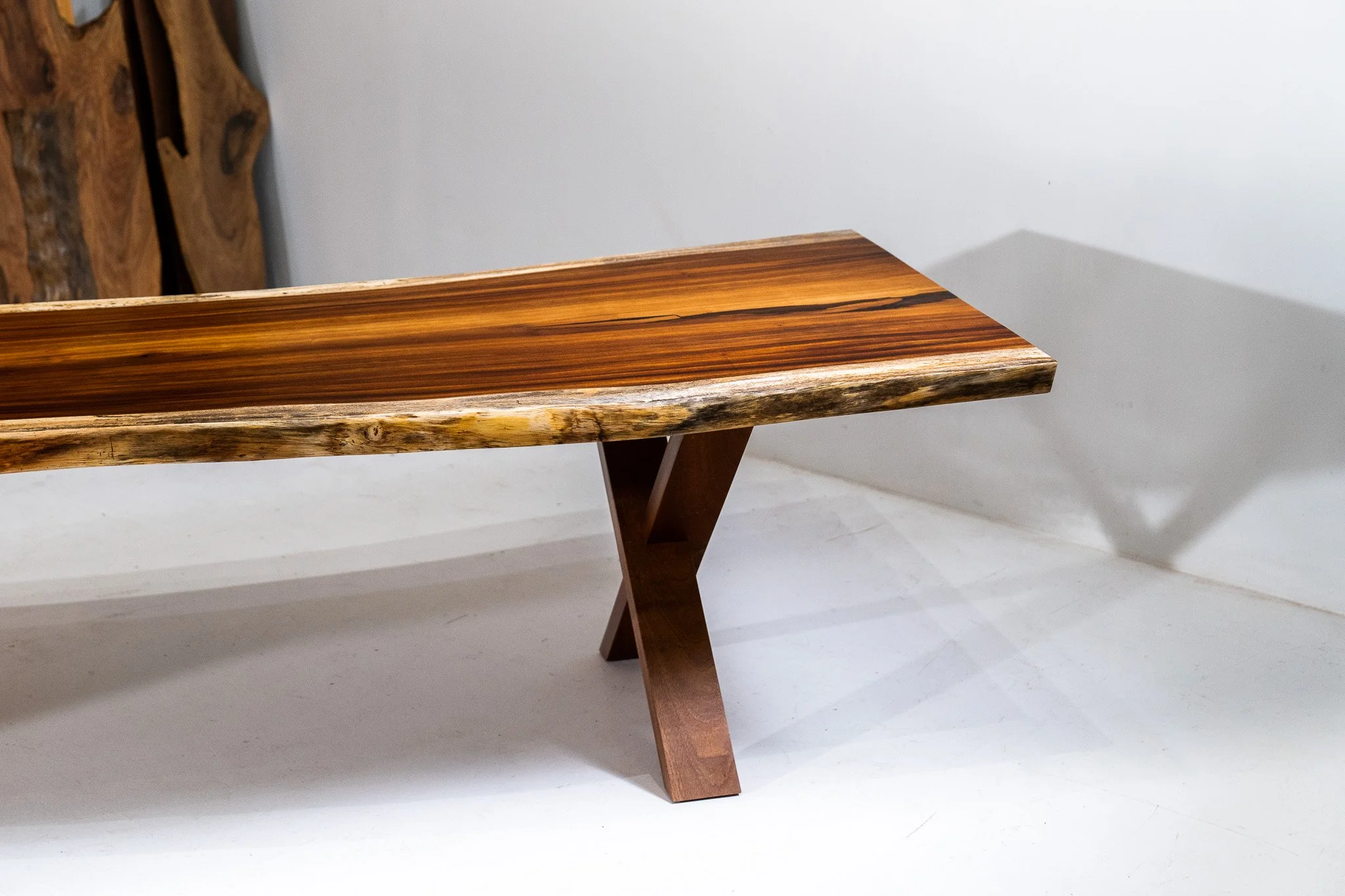 A wooden bench with a polished, dark wood seat and a natural, rougher wood edge, supported by tapered wooden legs, casting a shadow on a white background.