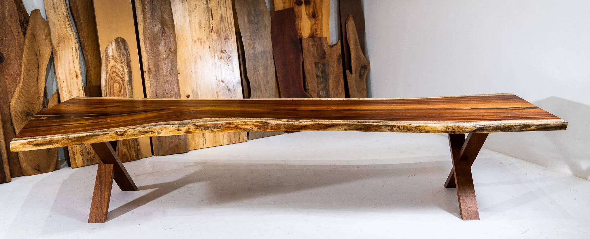 A wooden bench with a polished, live-edge surface and angled legs, placed on a white floor against a background of raw wood slabs leaning against a wall.