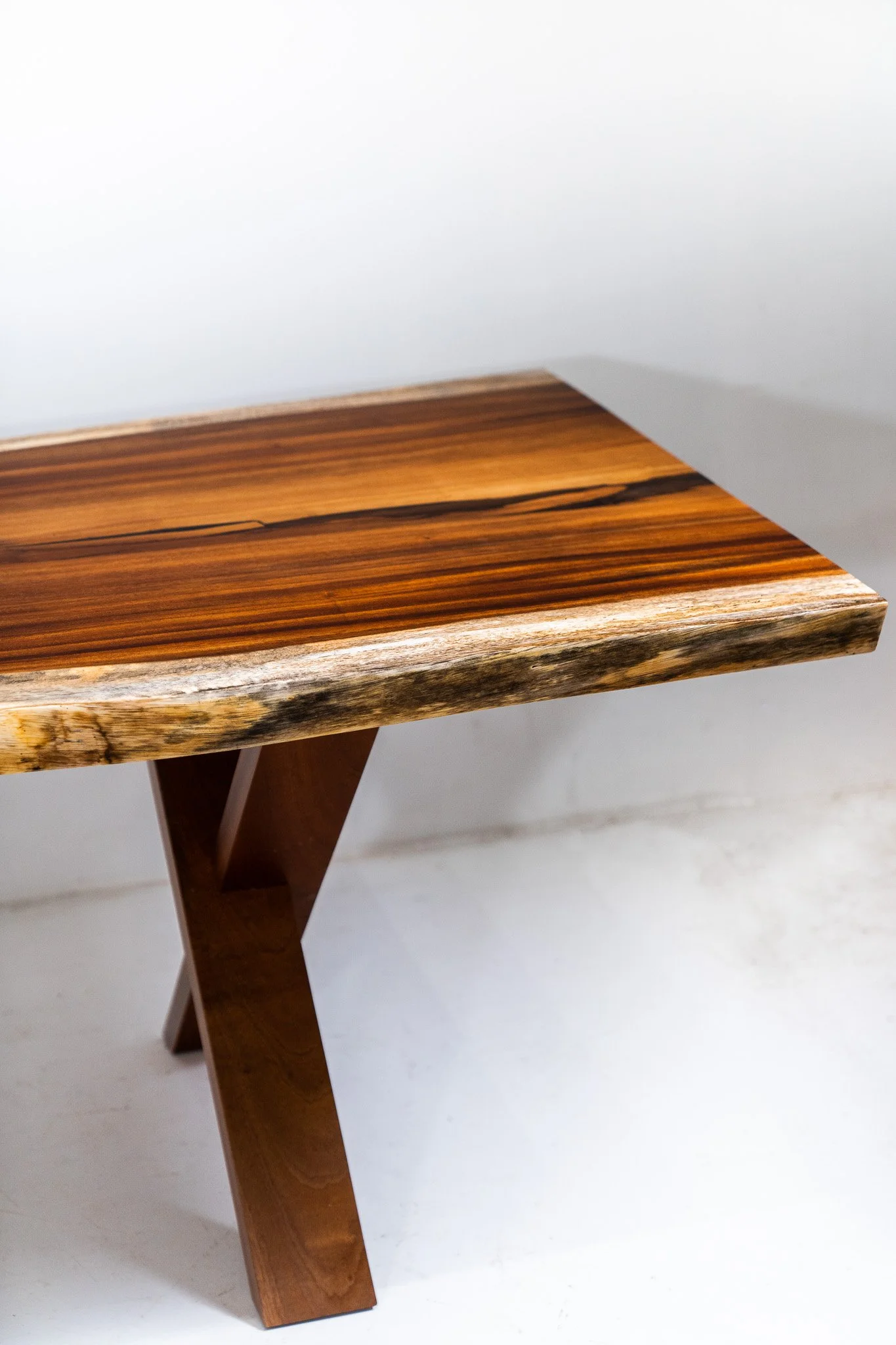 Close-up of a wooden table with a smooth polished top and a natural edge, supported by angled wooden legs, against a plain white background.