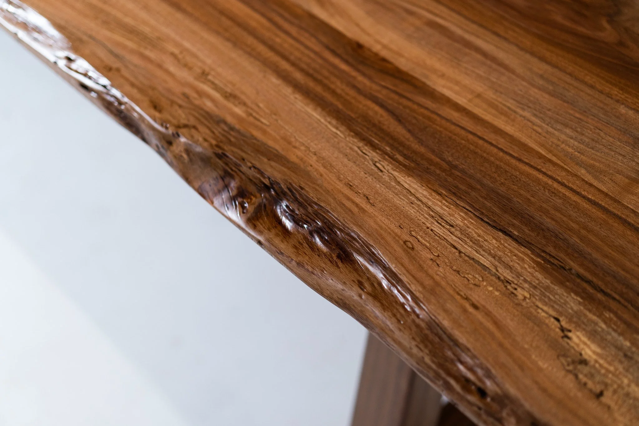 Close-up of a wooden table edge with a polished finish, showing natural wood grain and a live edge with uneven texture.